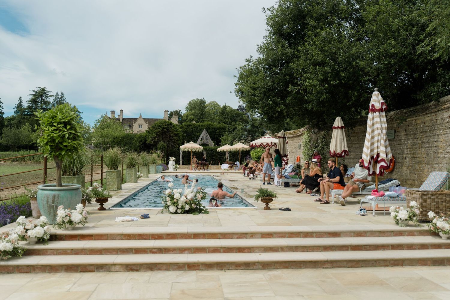 Wedding guests relax and mingle around an elegant outdoor pool at a countryside estate venue.