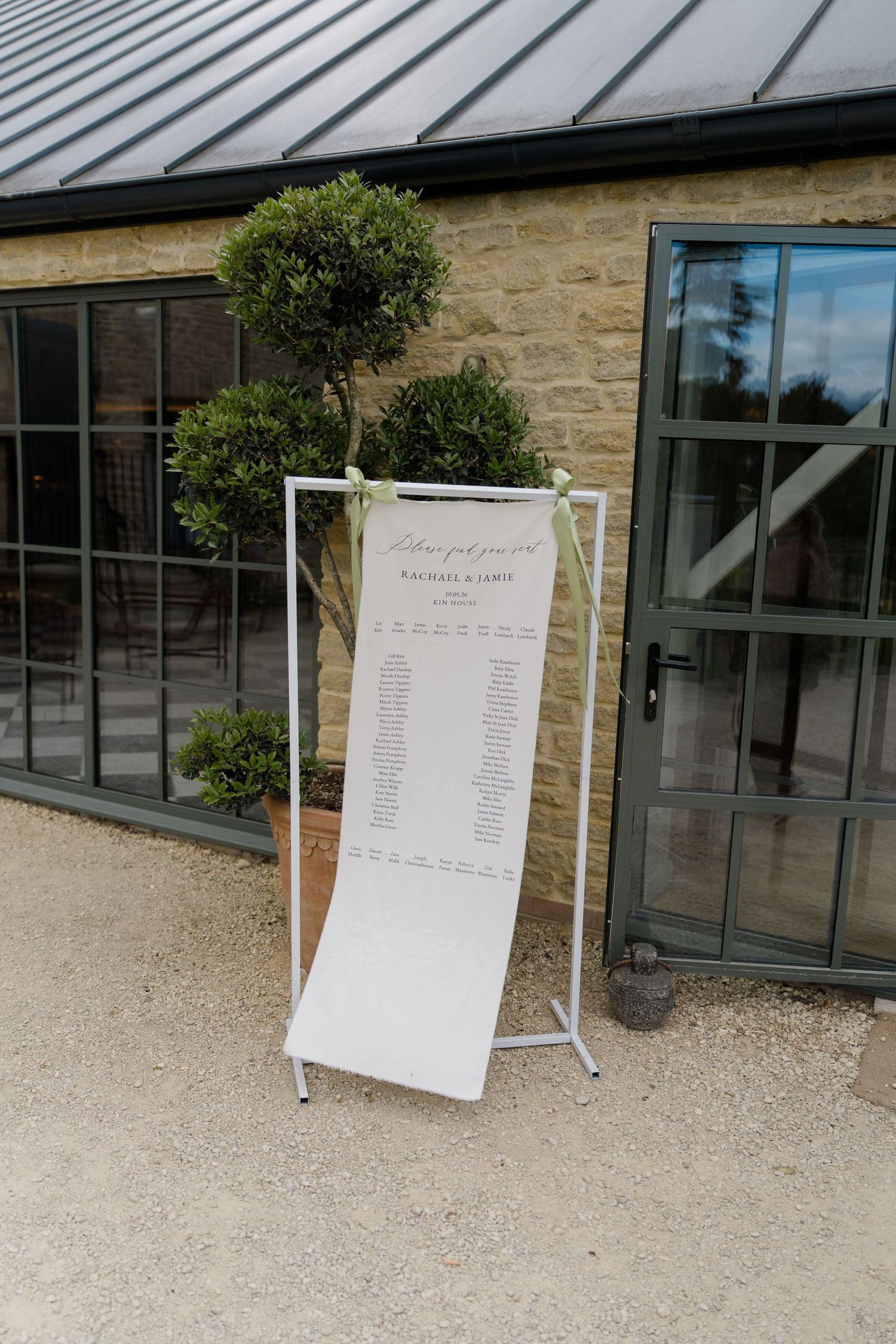 Elegant white seating chart on minimalist stand flanked by topiary trees outside stone venue.