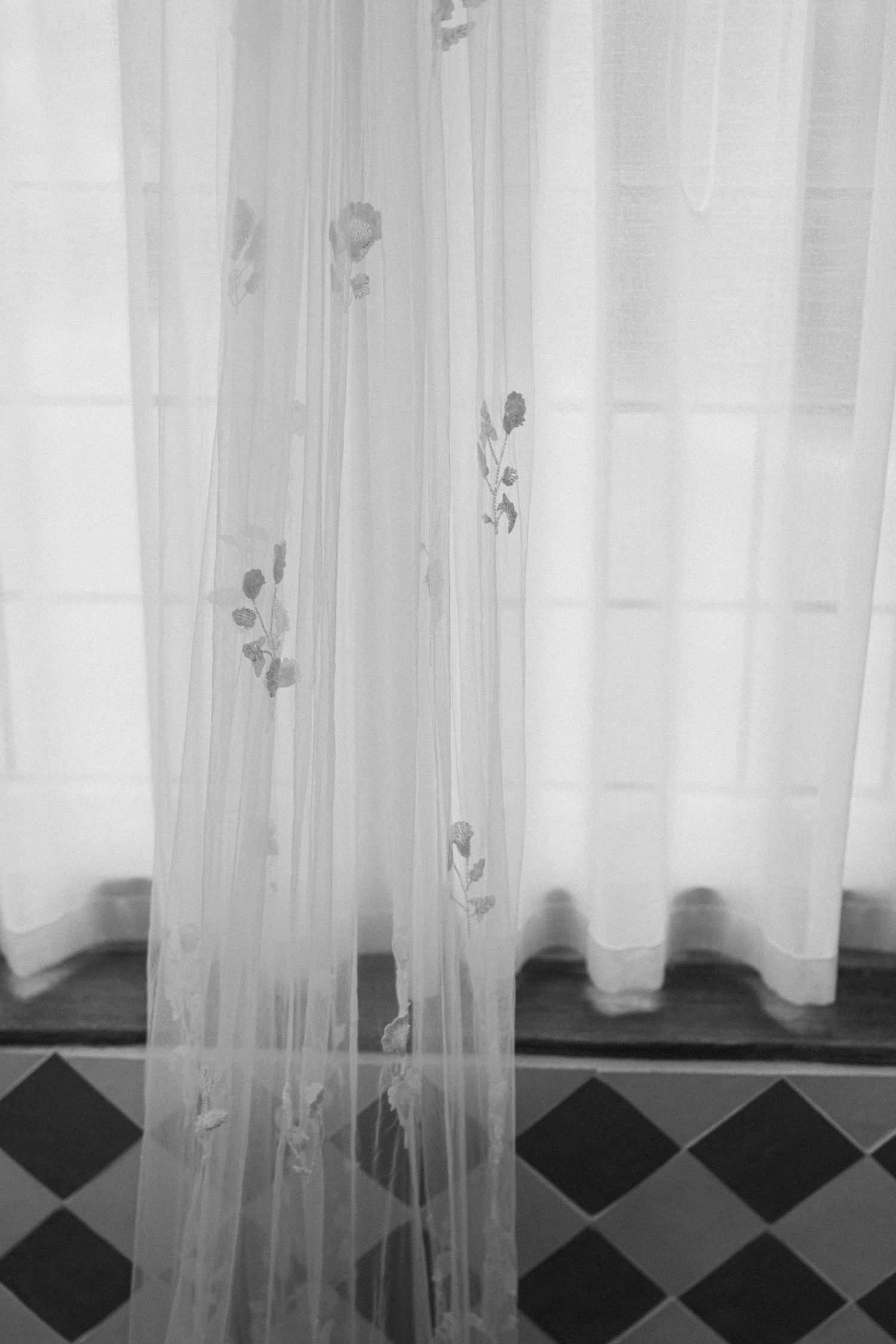 Black and white photo of sheer floral embroidered curtains over a black and white diamond tile floor.