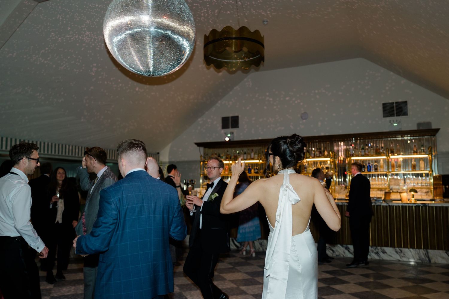 Bride and groom dance together under glowing disco ball near bar at lively wedding reception.