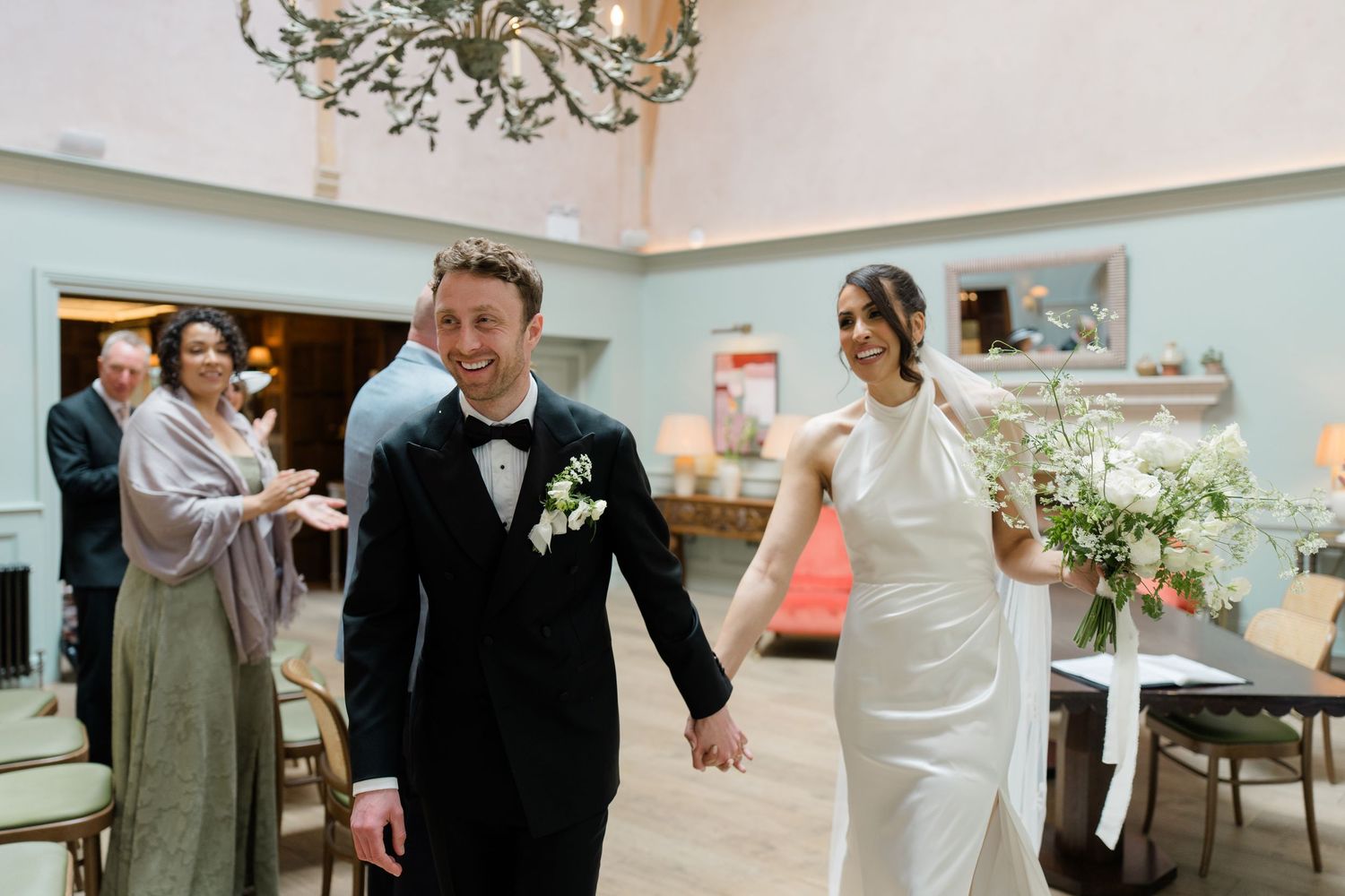 Newlywed couple walks hand in hand smiling after wedding ceremony, groom in black tuxedo, bride in white halter gown with bouquet.