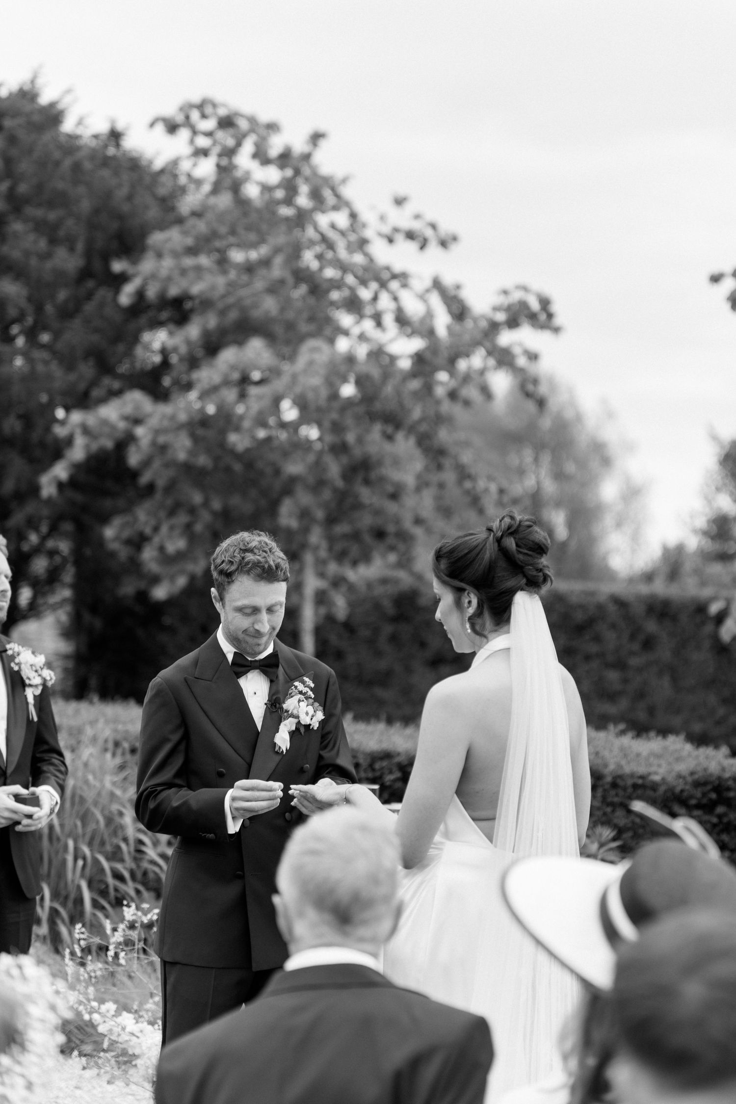 Groom placing wedding ring on brides finger during outdoor garden ceremony in black and white.
