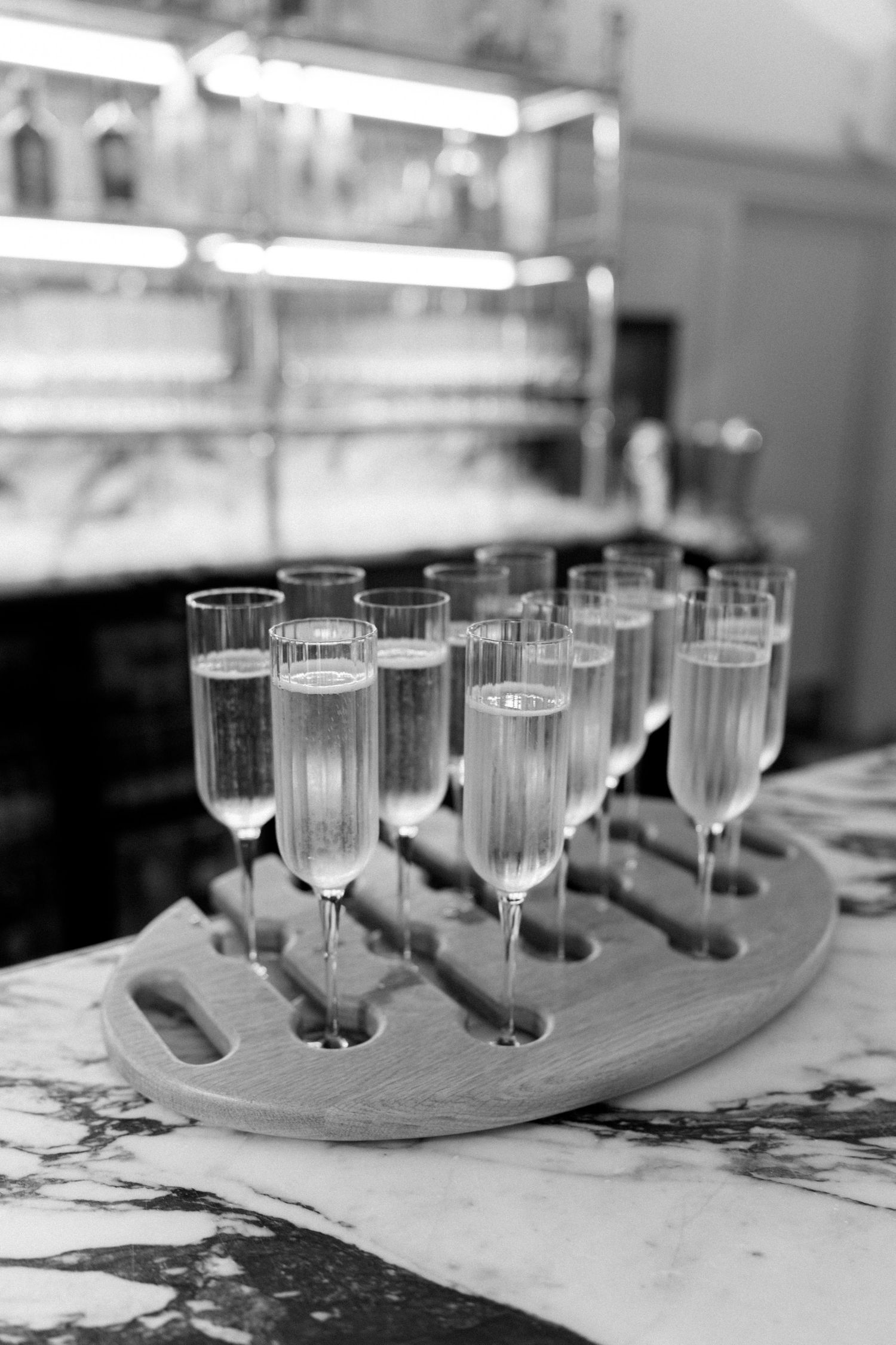 Black and white photo of champagne flutes arranged on a wooden tray atop a marble bar counter.