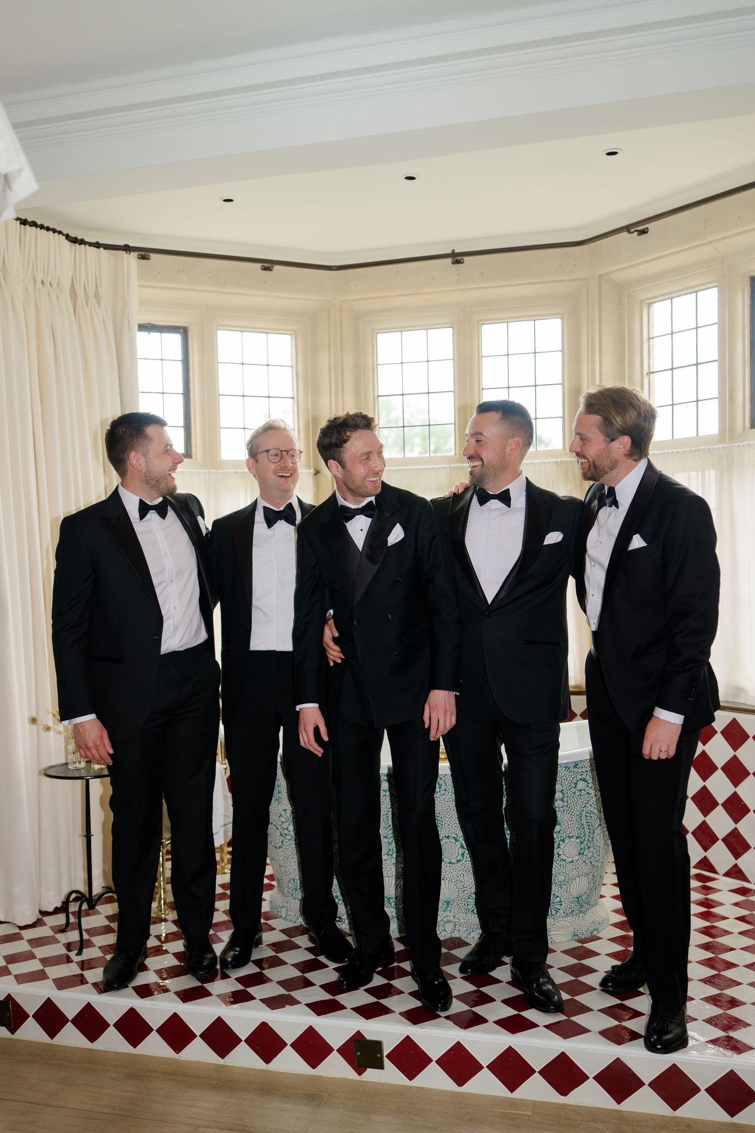 Five groomsmen in black tuxedos laughing together in elegant room with checkered floor.