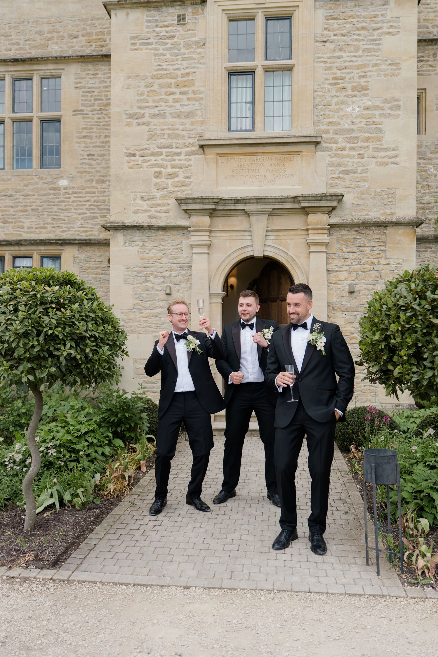 Three groomsmen in black tuxedos holding champagne glasses outside a grand stone manor building.