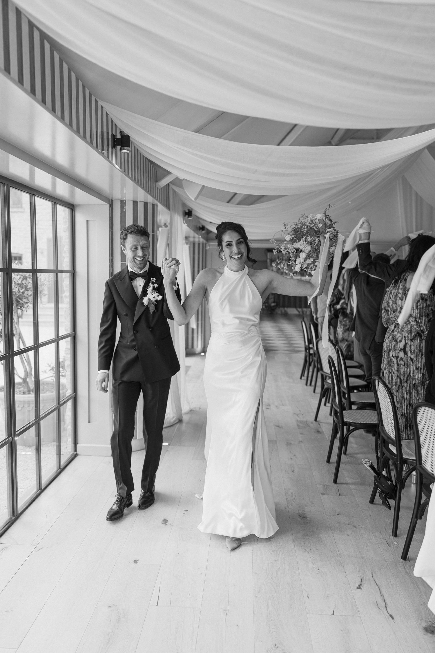 Joyful bride in white halter gown raises arm as groom in tuxedo enters decorated wedding reception.