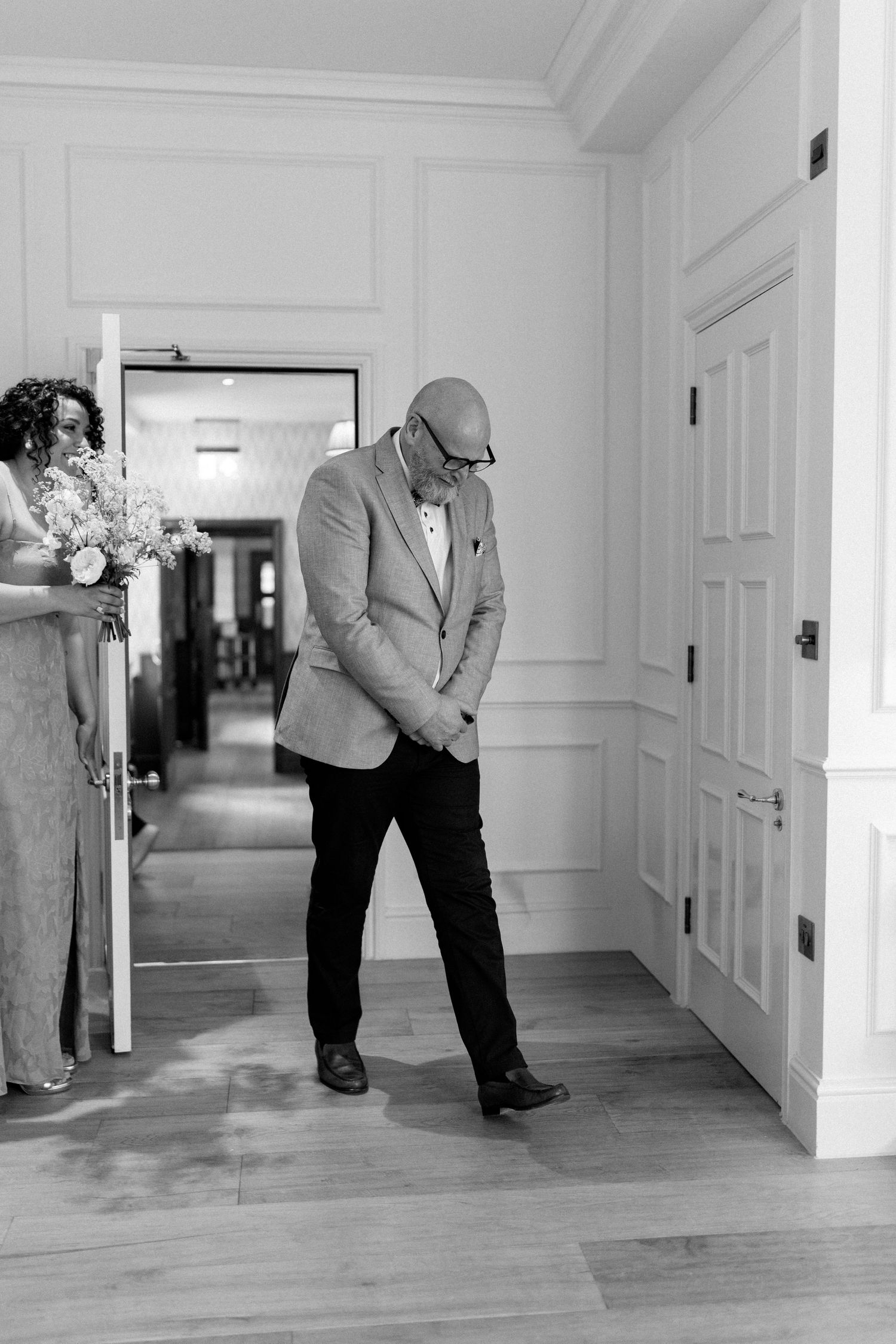 Emotional father bowing head in hallway as bride with bouquet approaches in background.