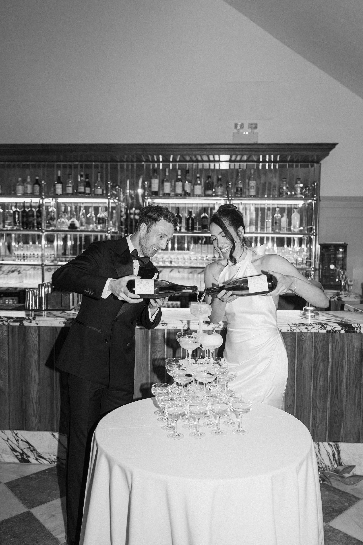 Bride and groom pour champagne tower together at wedding reception bar.