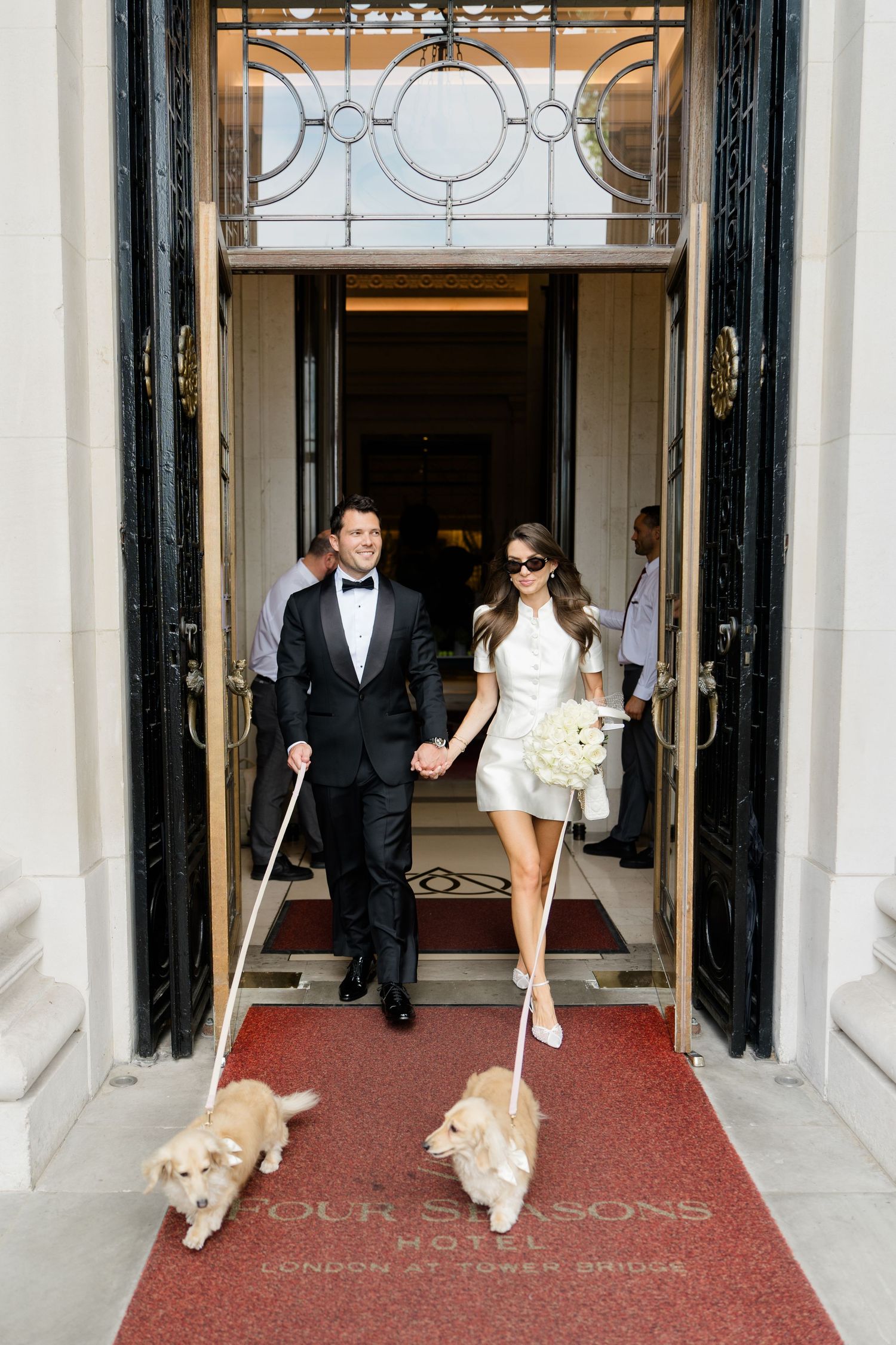 Newlyweds in wedding attire exit Four Seasons Hotel London with two golden dogs on leashes.
