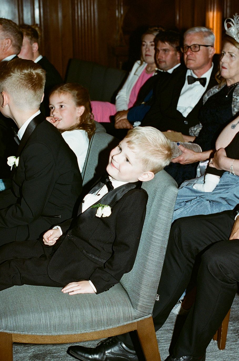 Children and adults in formal attire seated at a wedding ceremony, boys wearing black suits with white boutonnieres.