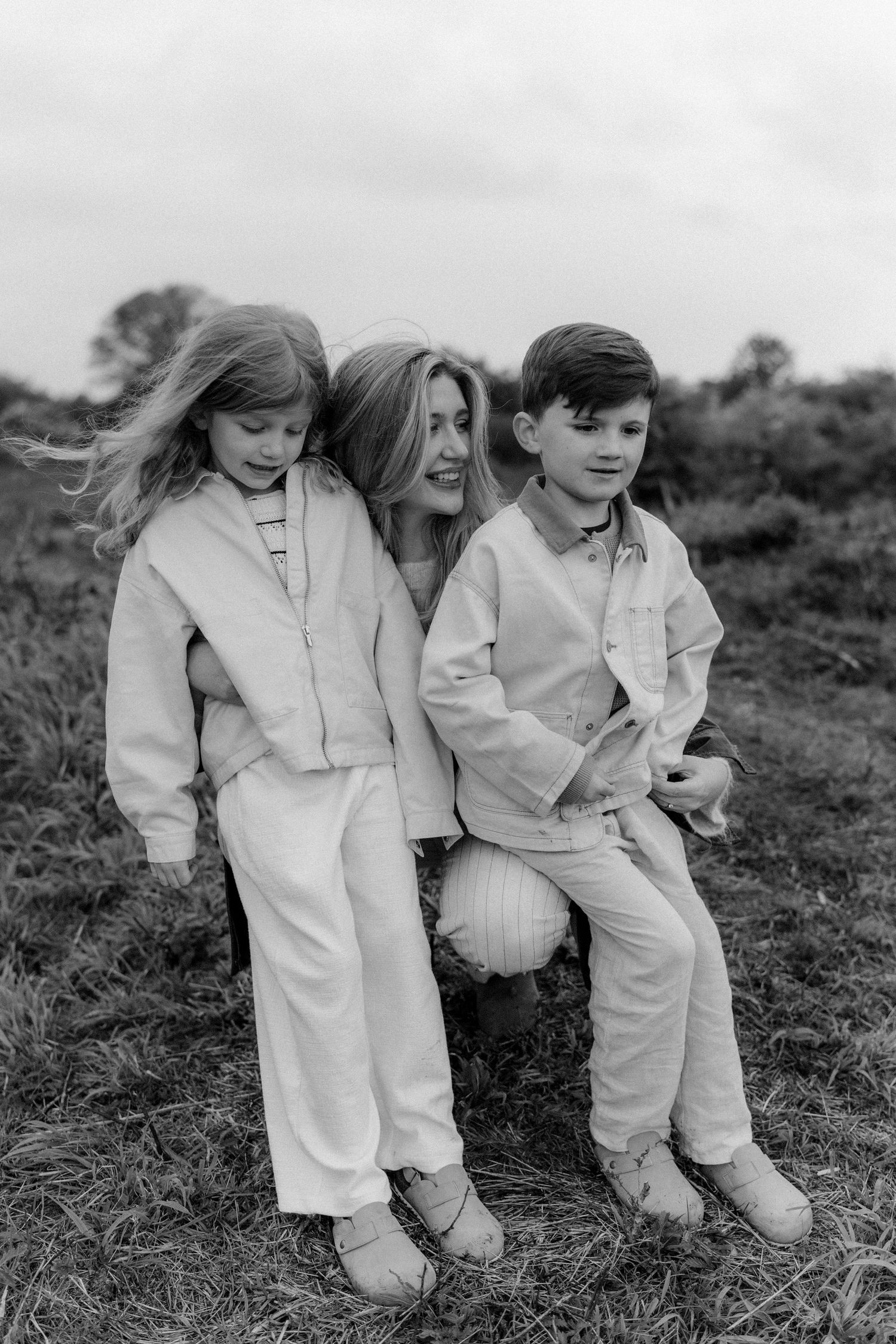 Black and white photo of a mother sitting outdoors with her two young children, a girl and a boy, in a grassy field.