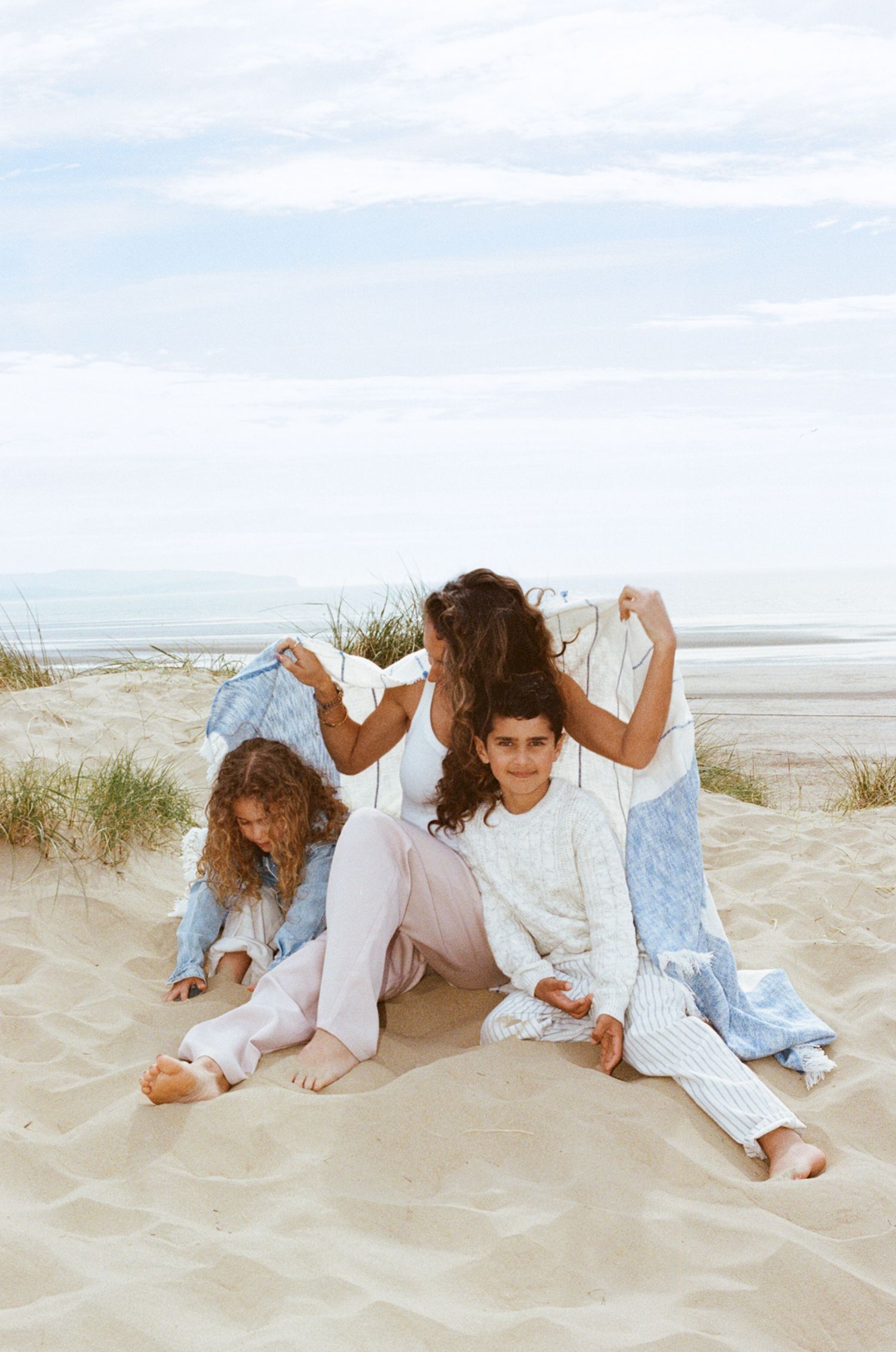 Mother and two children playing with blankets on sandy beach dunes under a cloudy sky.