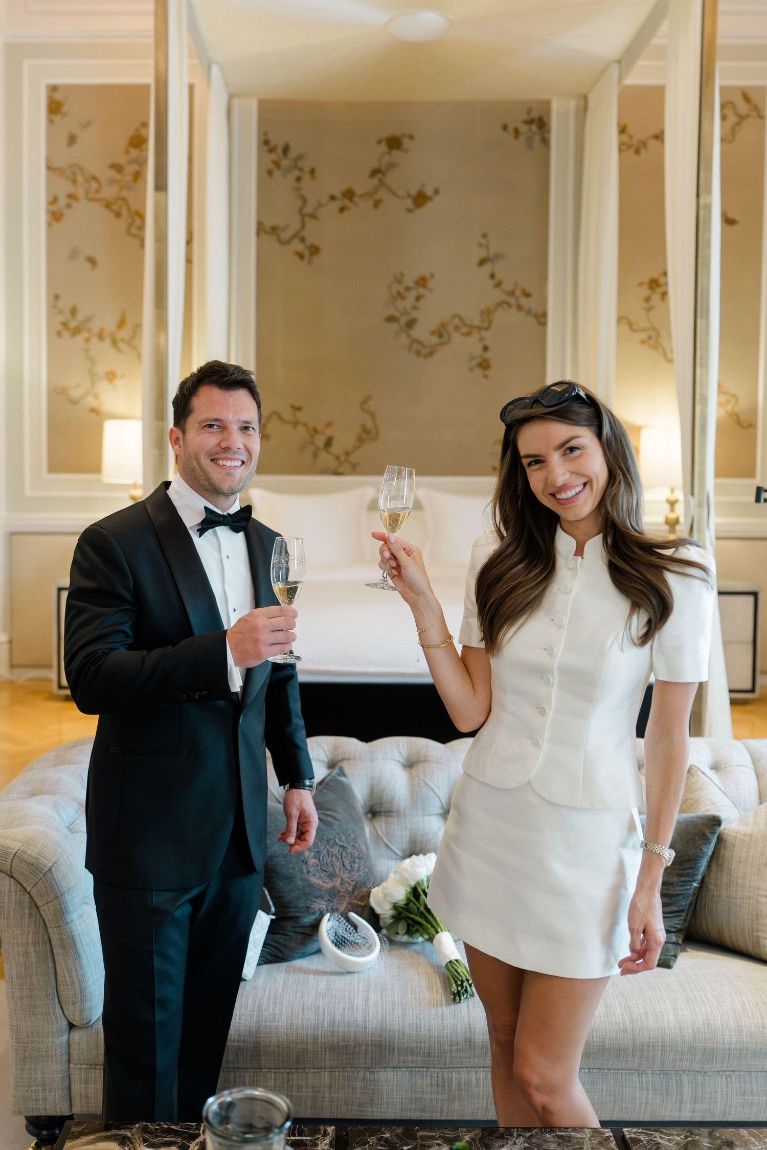 Elegant couple in formal attire toasting with champagne glasses in a luxurious hotel suite.
