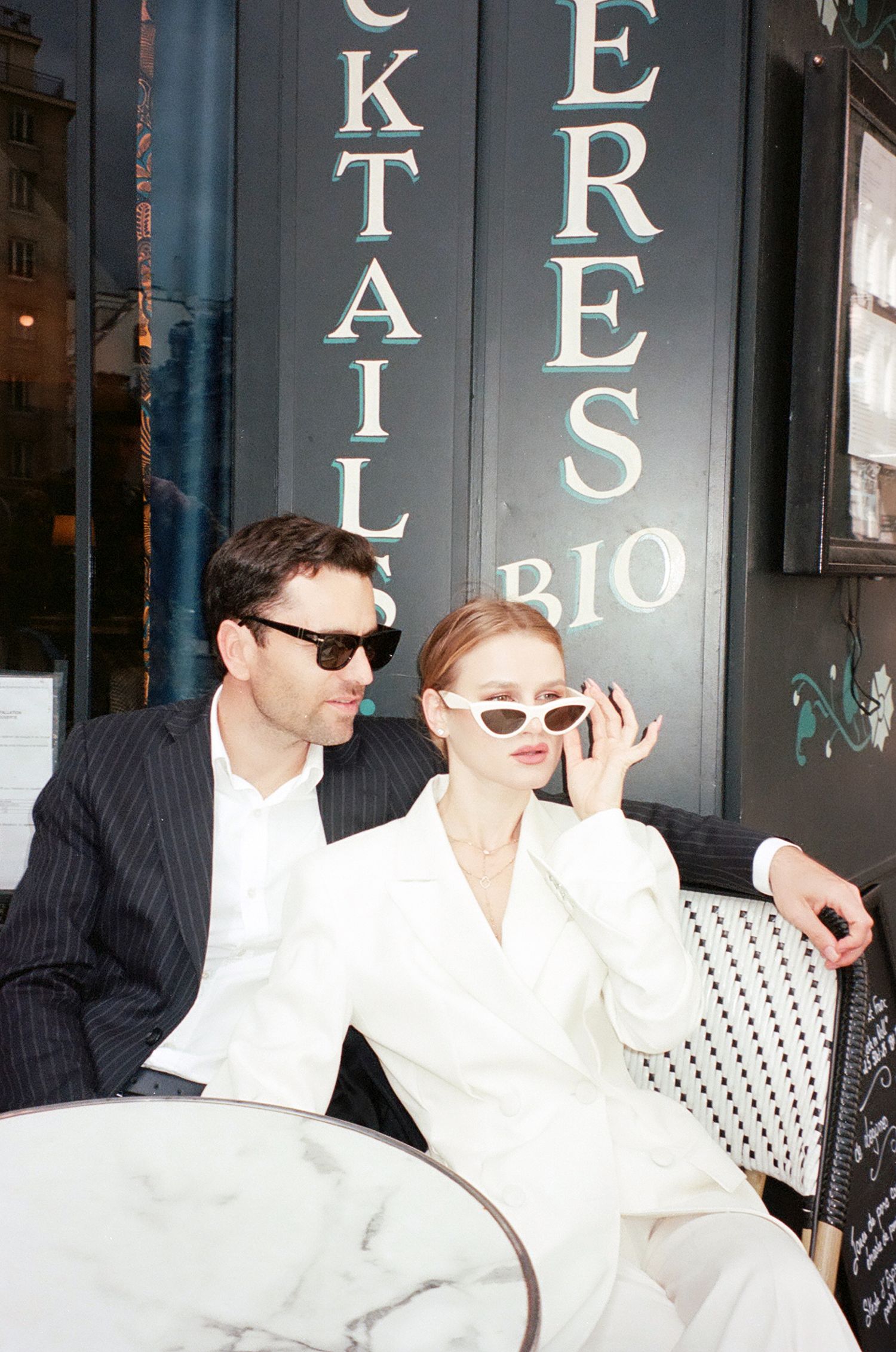 Three stylish figures in sunglasses sit together wearing black and white outfits outside a storefront.