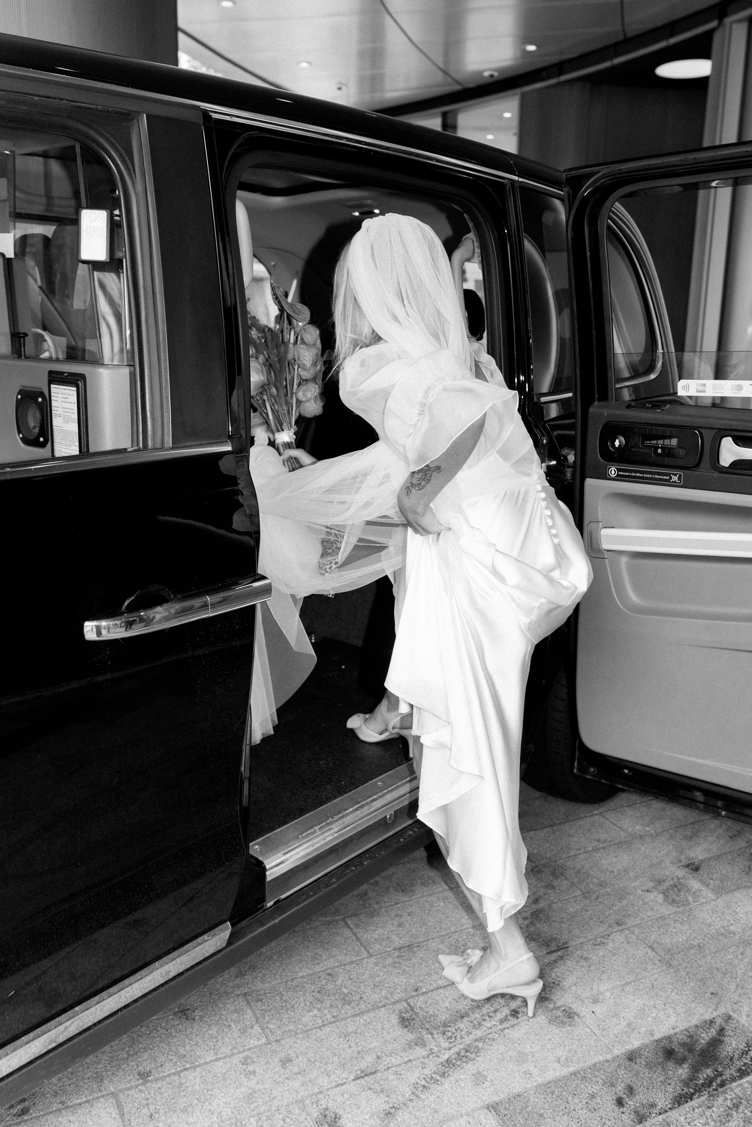 Black and white photo of bride entering vintage car while holding wedding dress train.