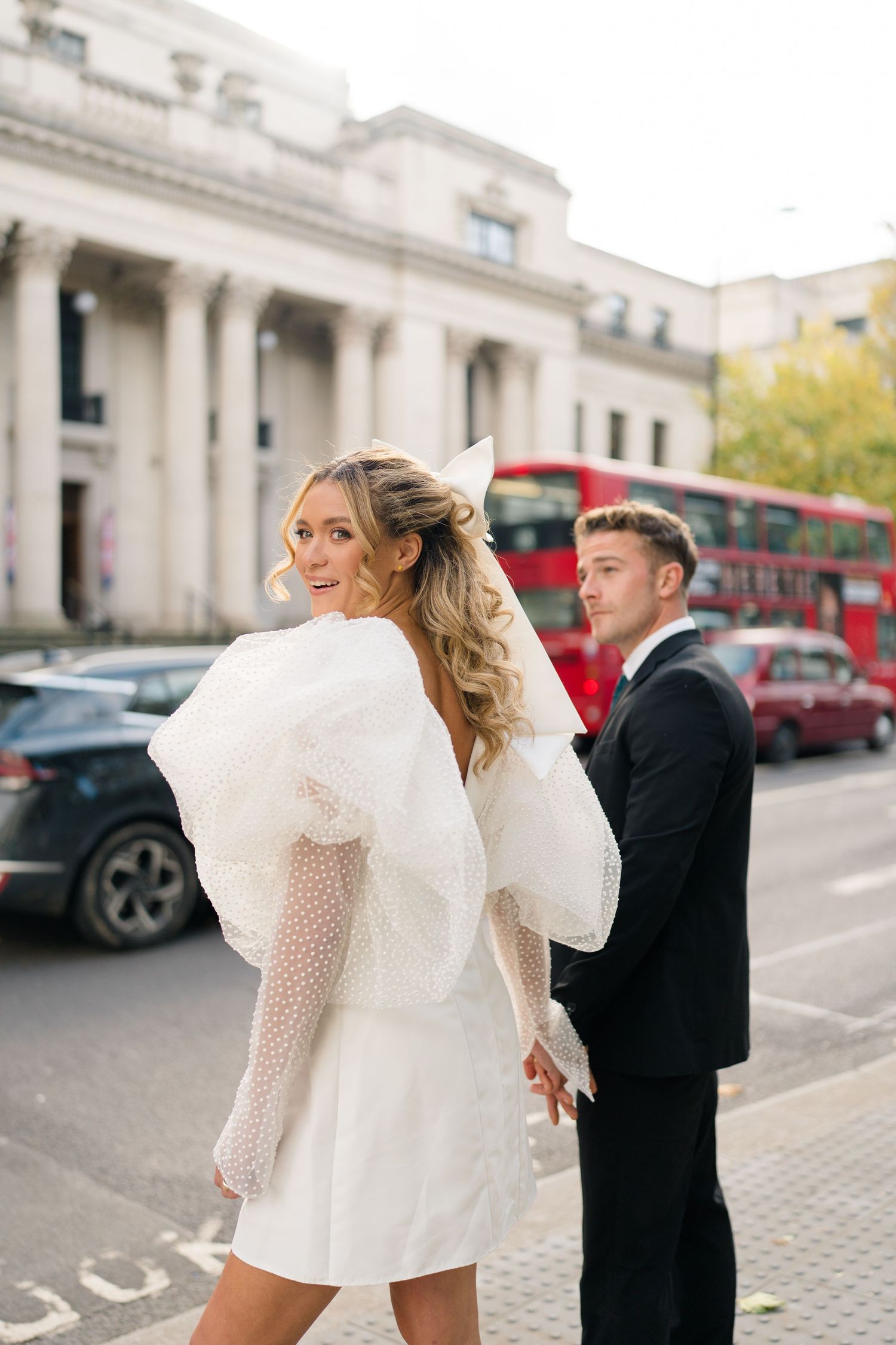 A couple in formal wear poses playfully on a London street with red buses visible.