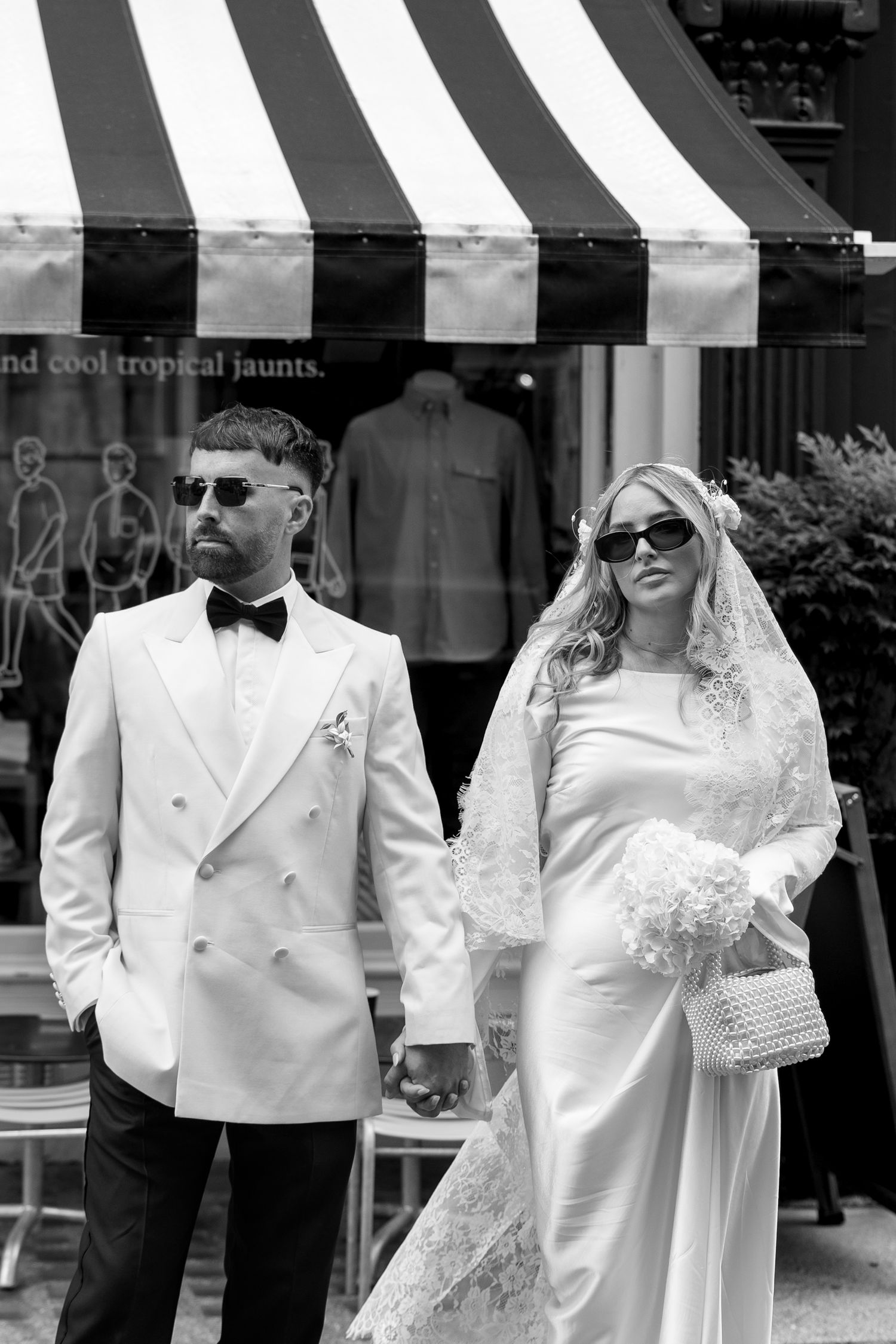 A wedding couple in white formal attire posing under a striped awning.