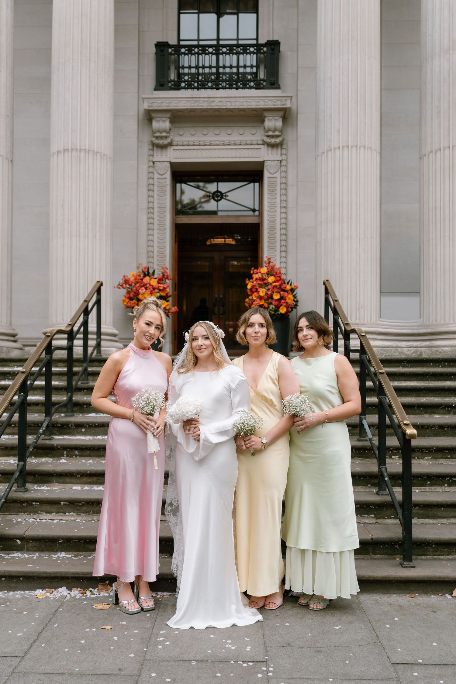 A bridal party in pastel dresses poses on grand stone steps with floral arrangements.