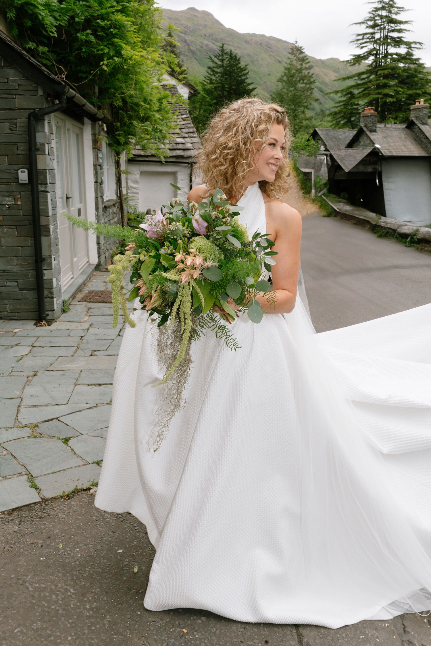 Bride in white wedding dress walking along stone path with mountains and greenery in background.