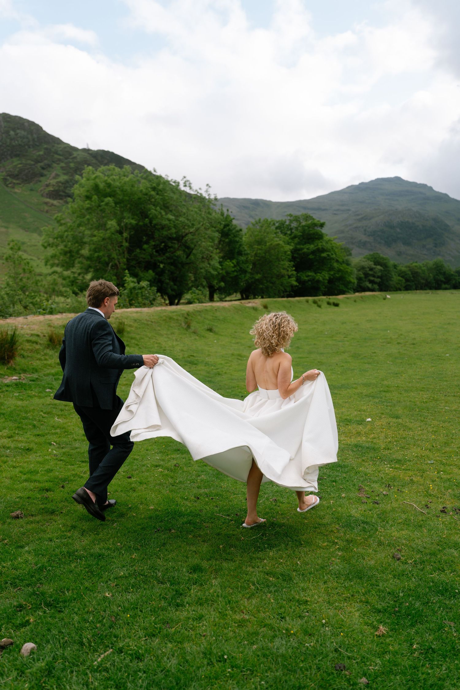 A couple playfully runs through a green meadow with mountains in the background during a wedding photoshoot.