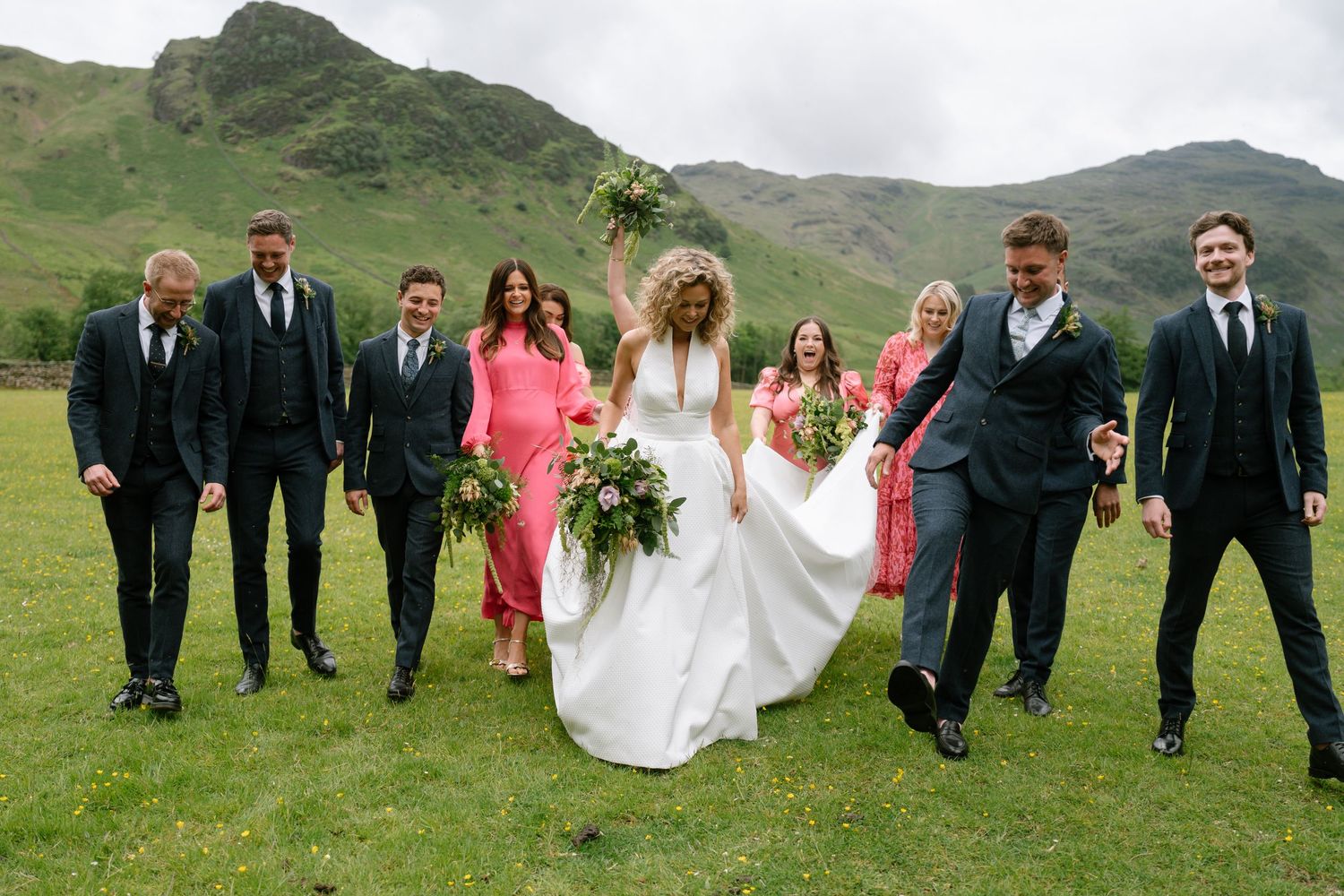 Wedding party walking in formation across green hillside with mountains in the background.