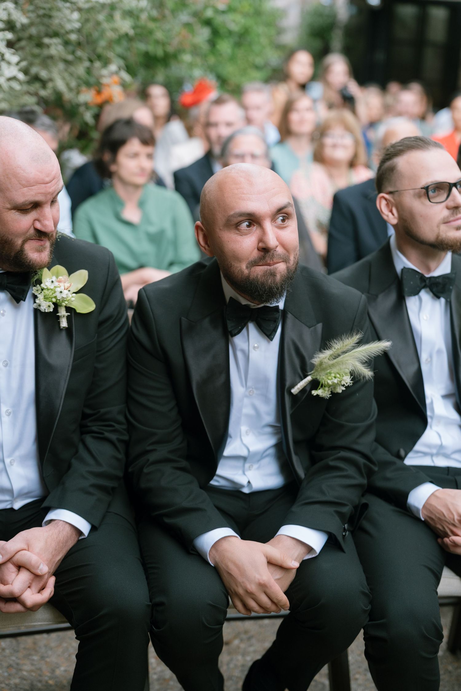 Groomsmen in black tuxedos and bow ties sit in a row at an outdoor wedding ceremony.
