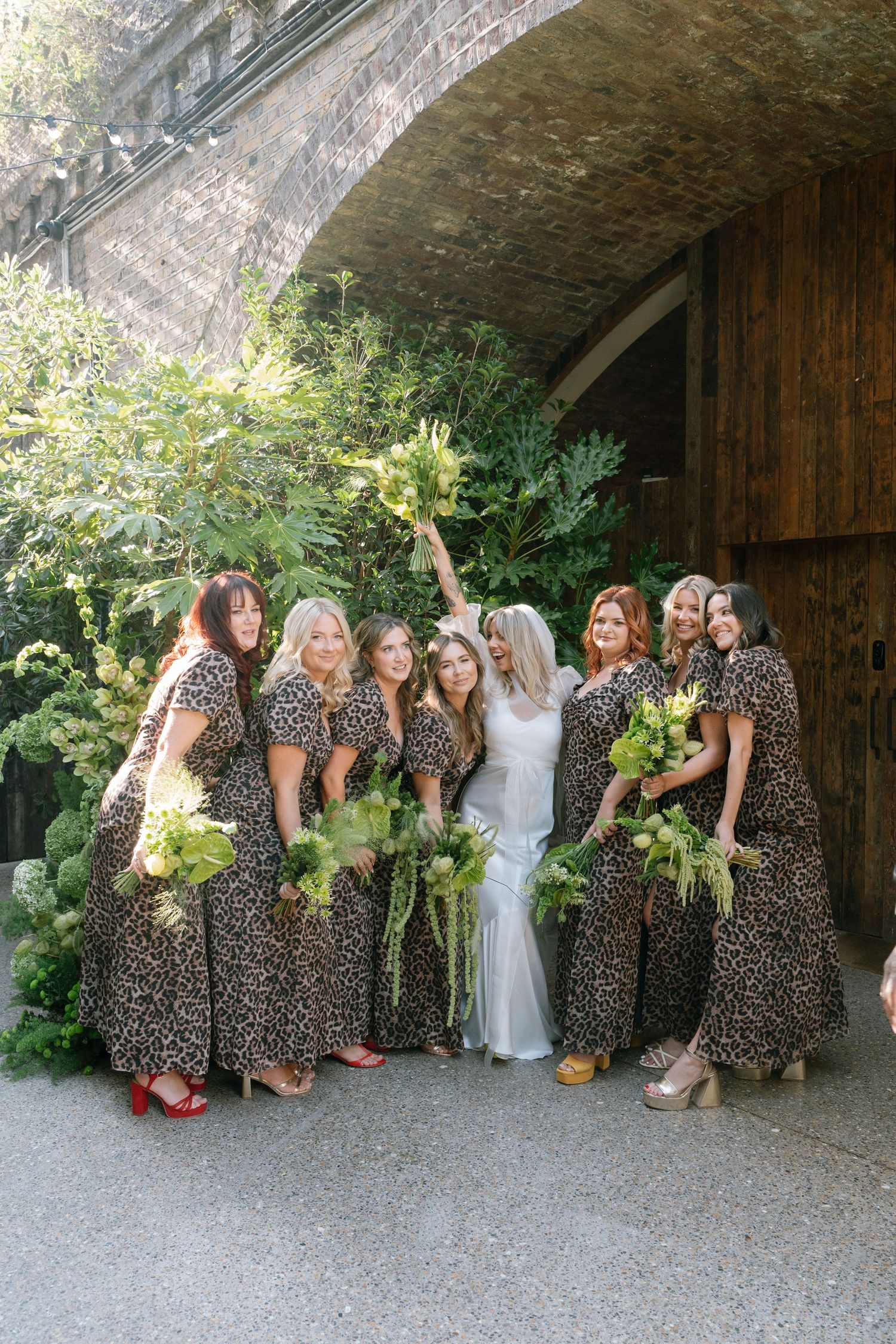 Wedding party wearing matching leopard print dresses pose together under a rustic archway with floral bouquets.
