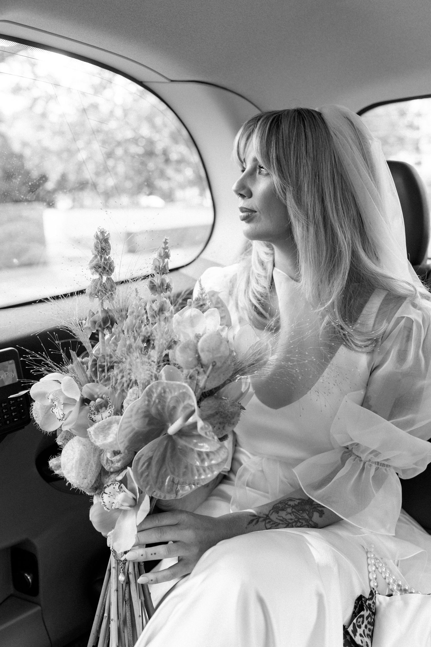 A bride in a white dress sits in a car holding a lush bouquet of flowers in black and white.