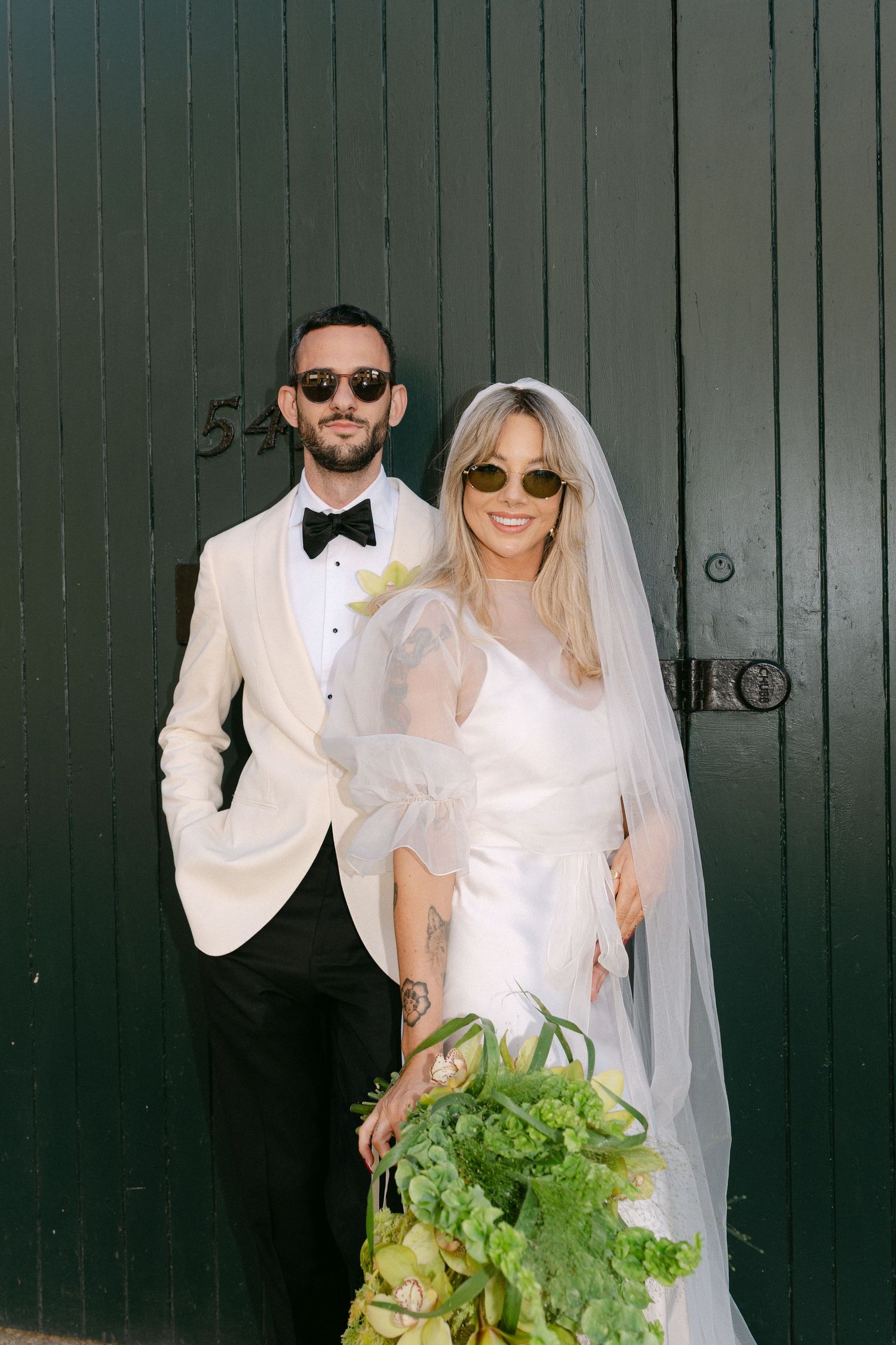 Newlyweds pose against green doors holding fresh lettuce bouquets.
