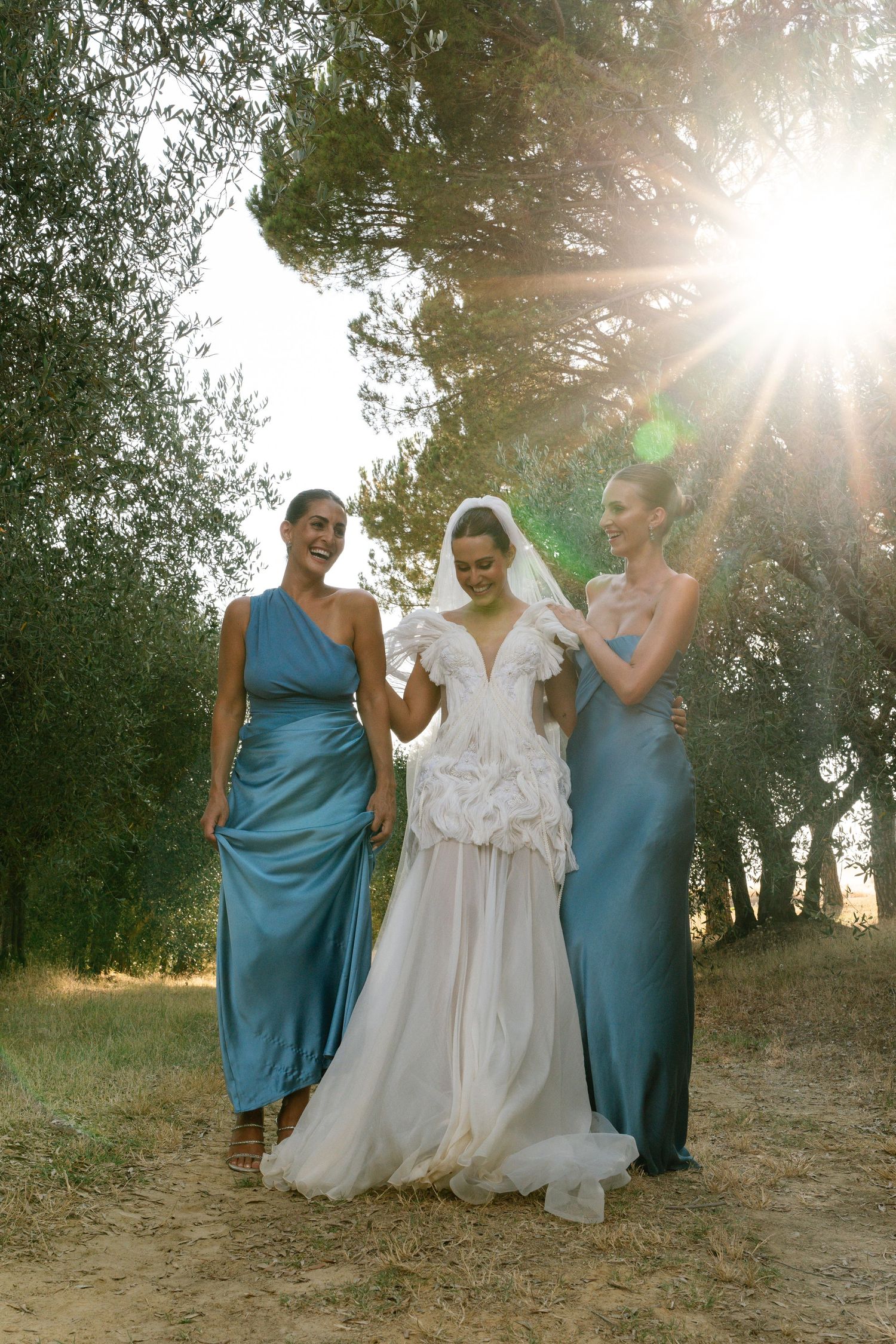 Three people in formal attire pose outdoors with sunlight streaming through trees, featuring blue dresses and a white gown.