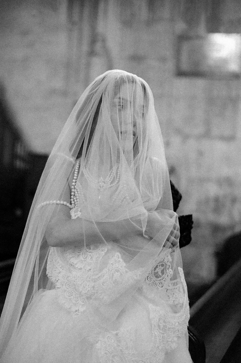 A bride in a delicate white lace wedding dress stands covered by a sheer veil in black and white photography.
