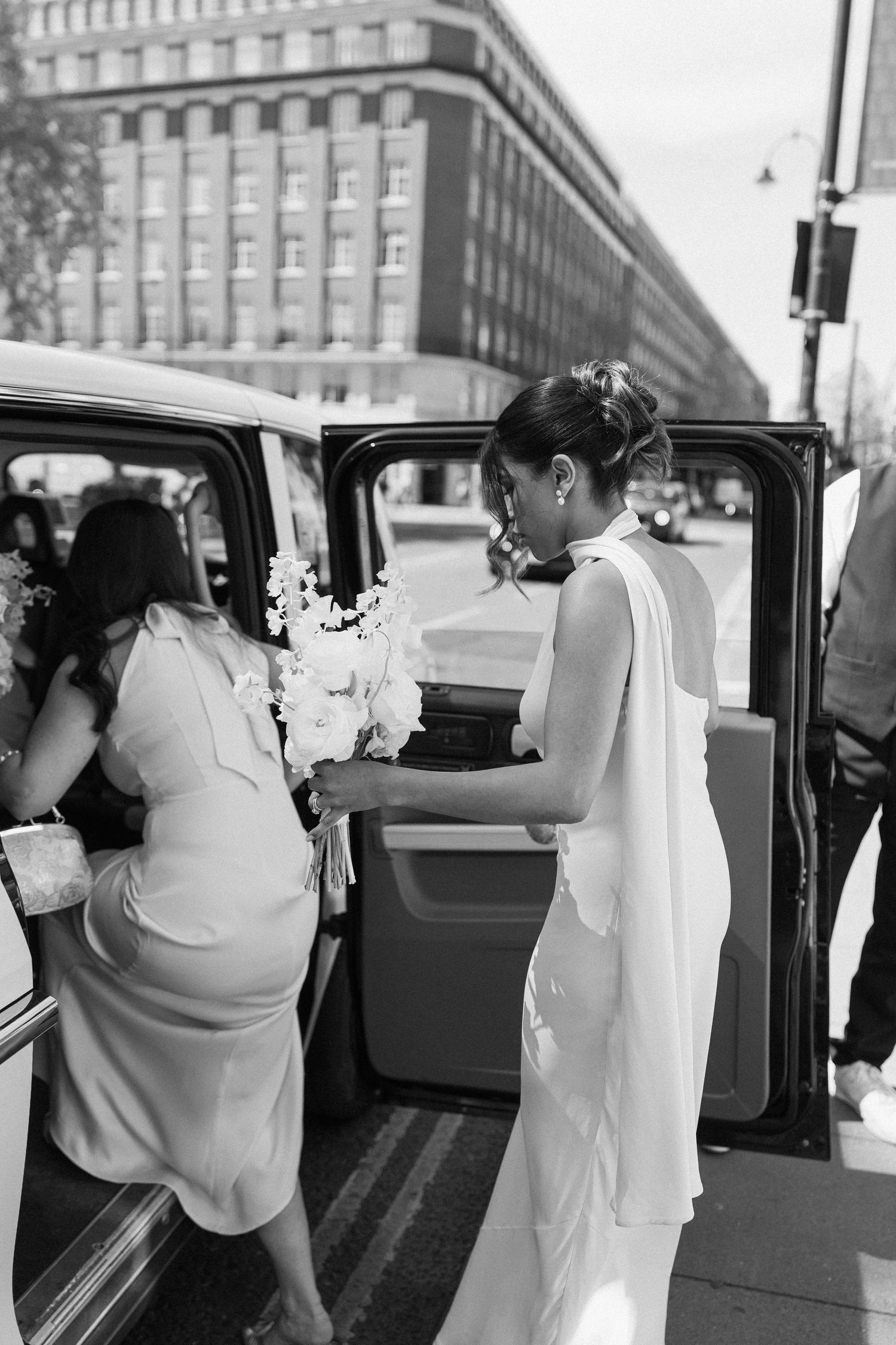 Bride in elegant white gown holding bouquet entering black taxi cab on city street, black and white.