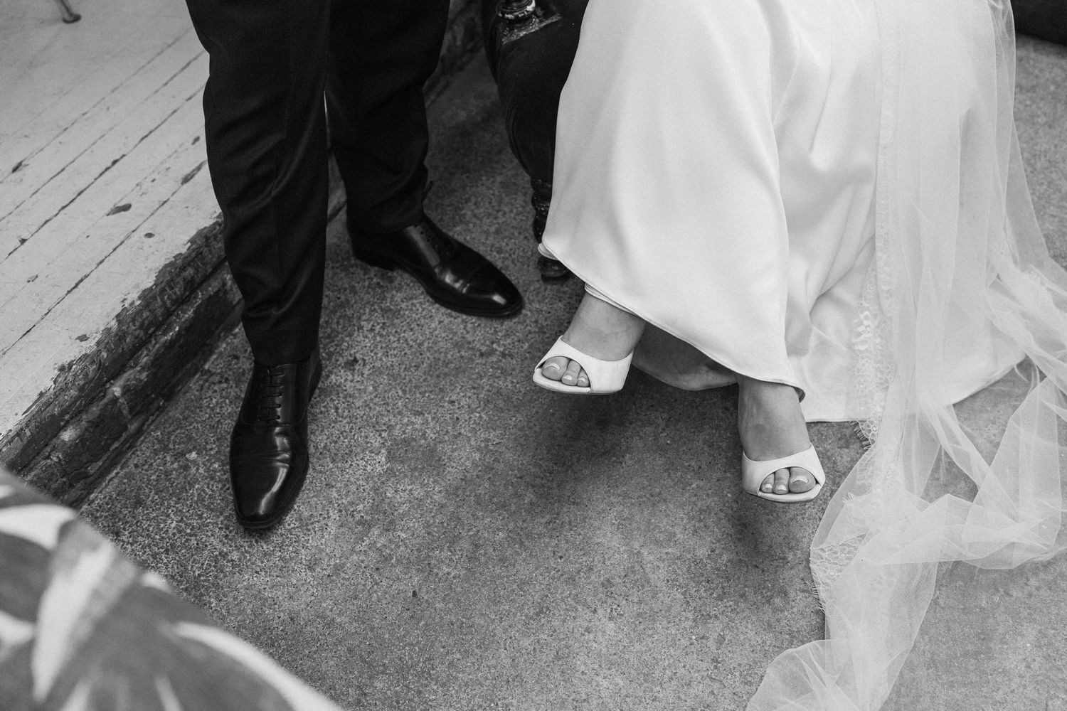Black and white wedding photo of bride in white gown and groom in black suit, focusing on their shoes on pavement.