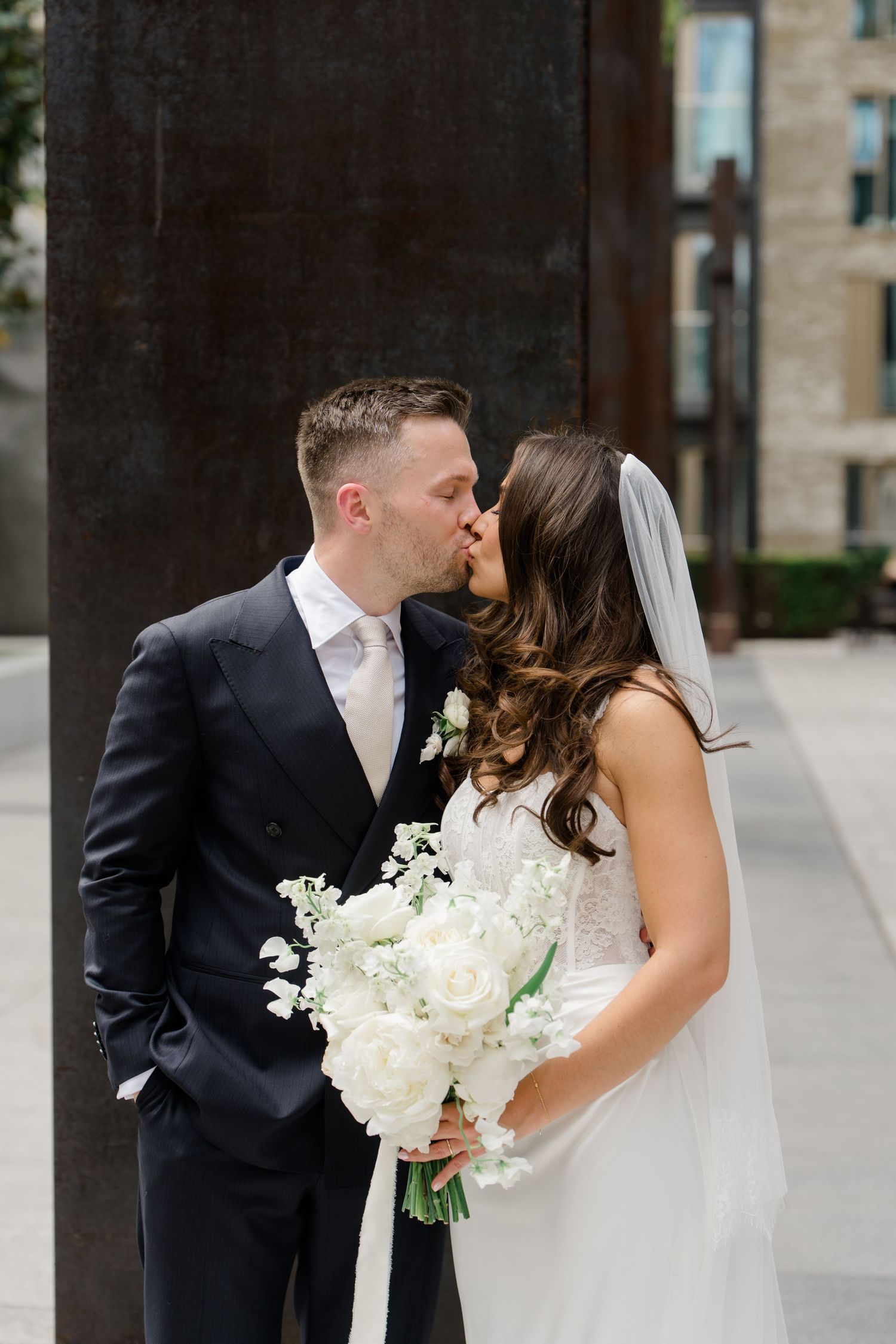 Bride and groom share a romantic kiss outdoors, bride holding a bouquet of white peonies and lily of the valley.