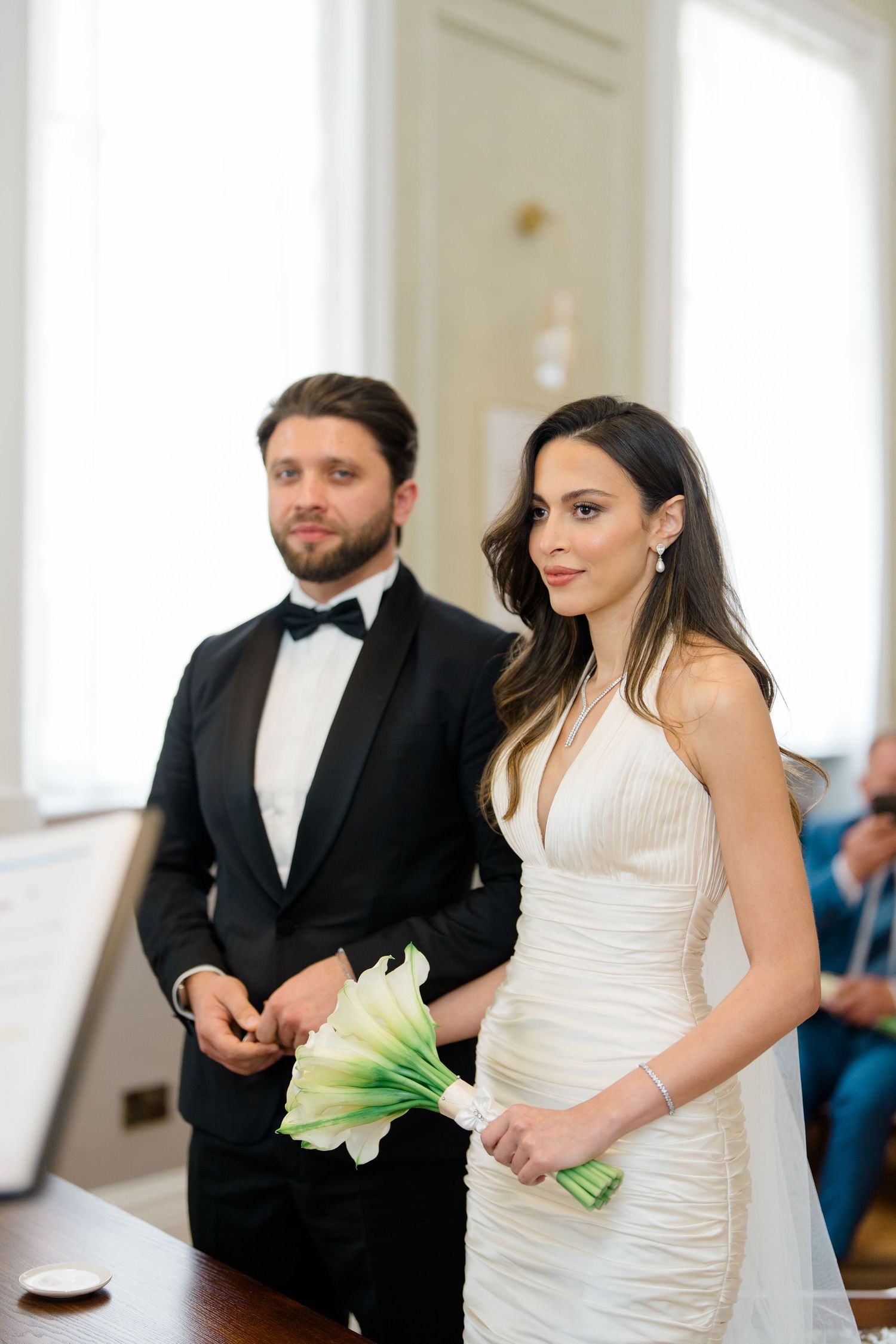 Bride in white halter gown holding calla lily bouquet stands with groom in black tuxedo during wedding ceremony.