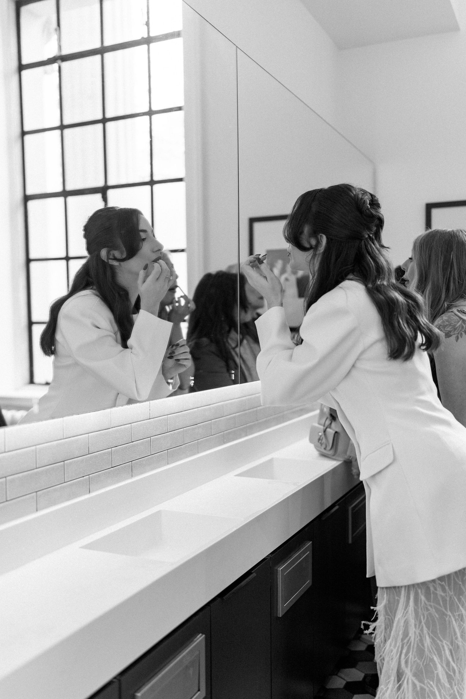 Black and white photo of a bride in a white blazer fixing her makeup at a bathroom mirror.