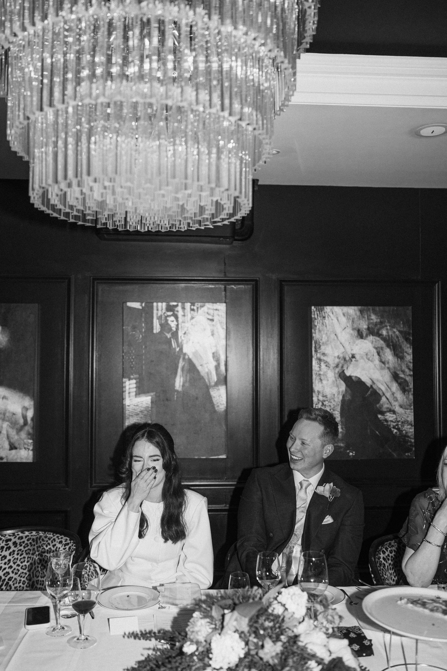 Black and white photo of laughing bride and groom seated at decorated wedding reception dinner table.