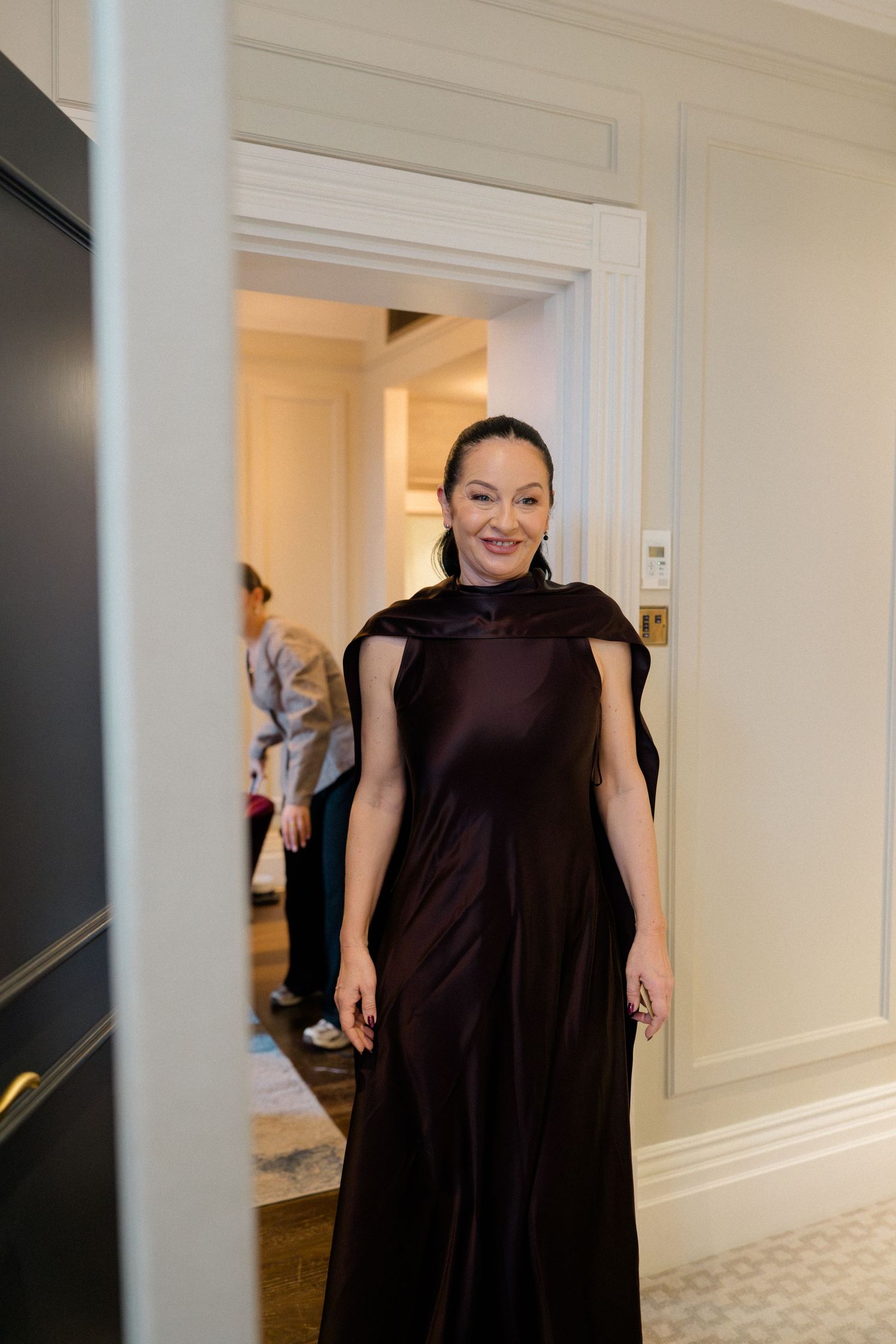 Woman in elegant dark burgundy satin cape gown smiling in a luxurious hotel hallway.