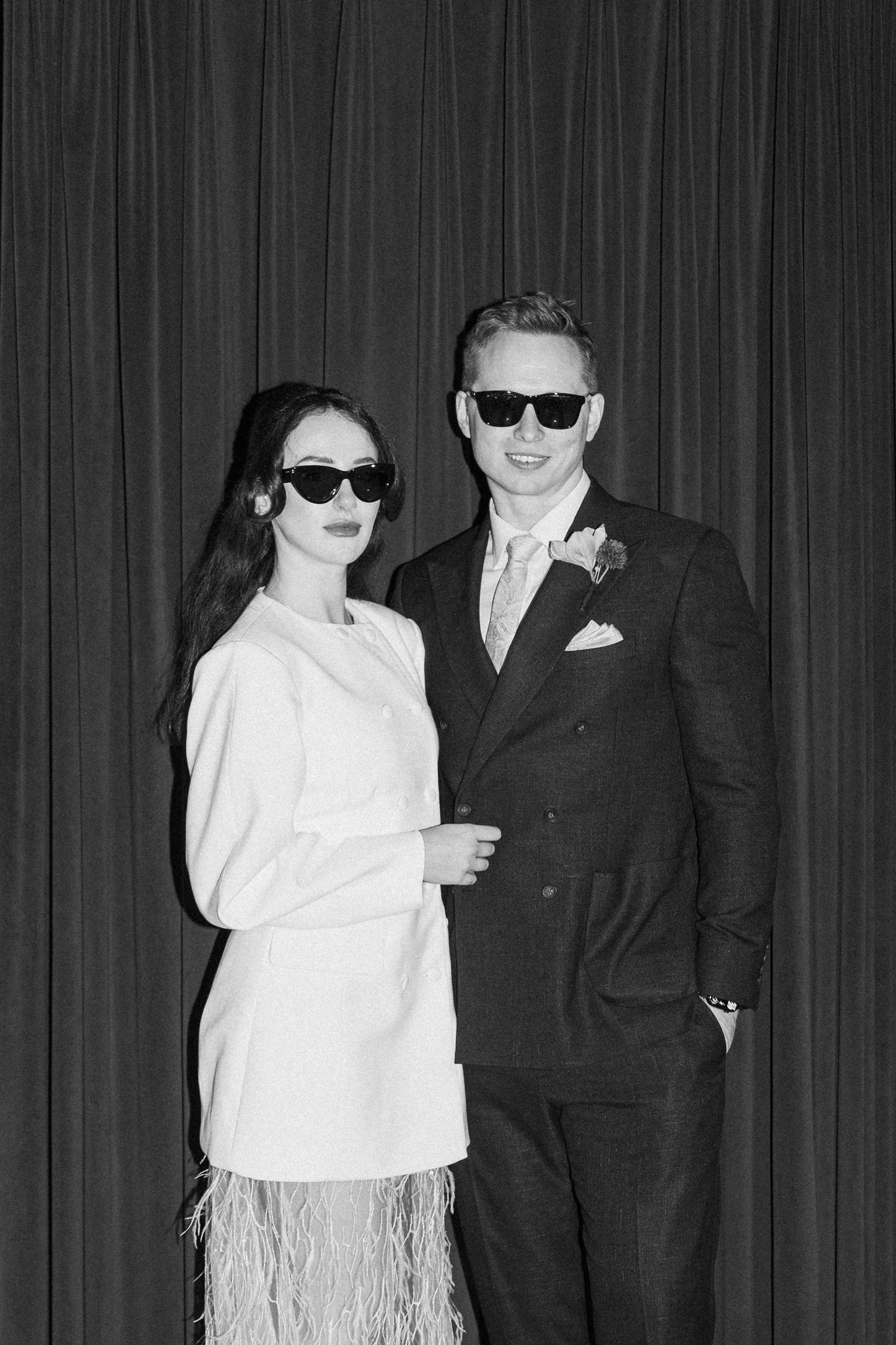 Stylish bride and groom pose in sunglasses wearing white mini dress and dark suit against draped curtain backdrop.