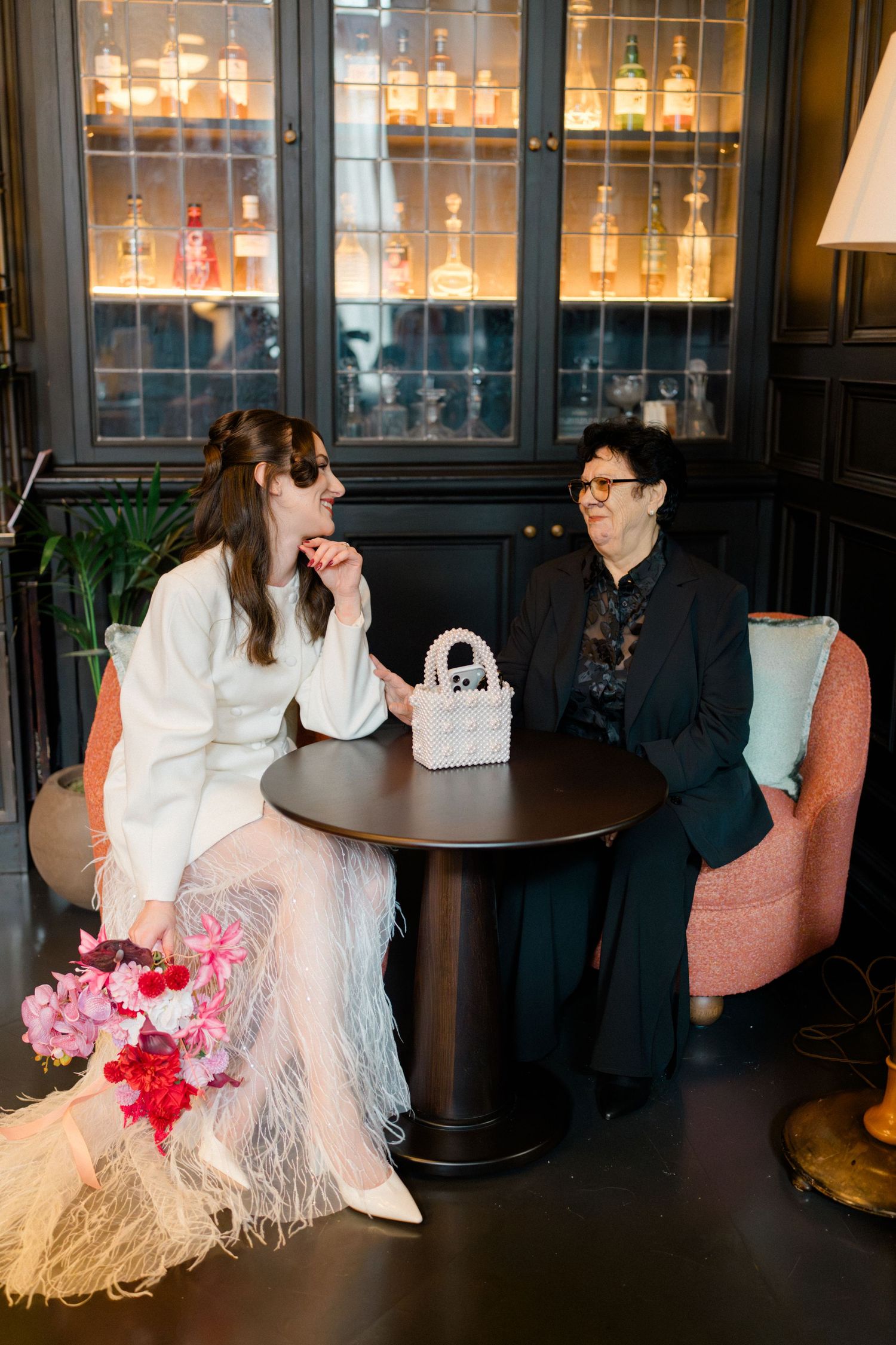 Bride in white feather skirt with sunglasses and pink bouquet chatting with guest at dark bar table.
