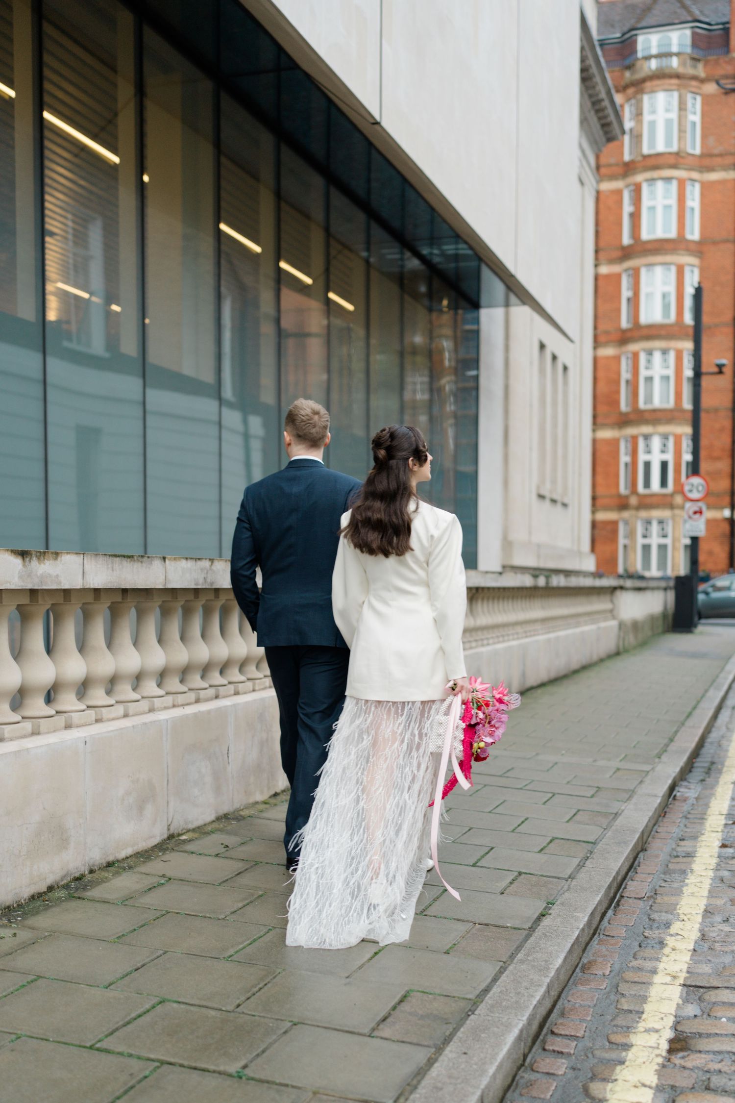 Bride and groom walking London street, bride in white blazer and feather skirt wedding outfit.