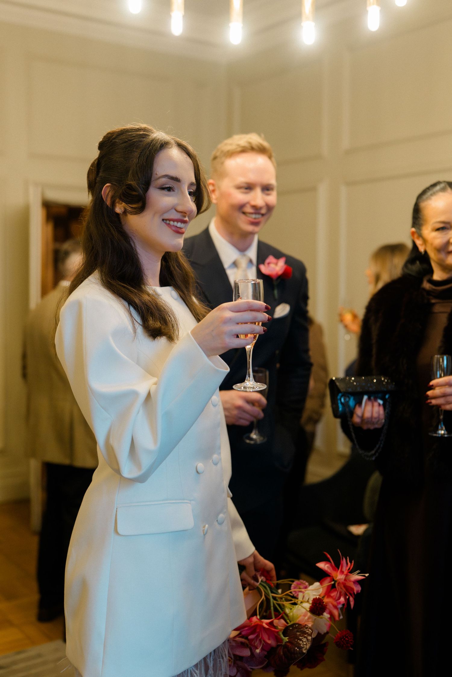 Smiling bride in white blazer holds champagne glass at intimate wedding reception celebration.
