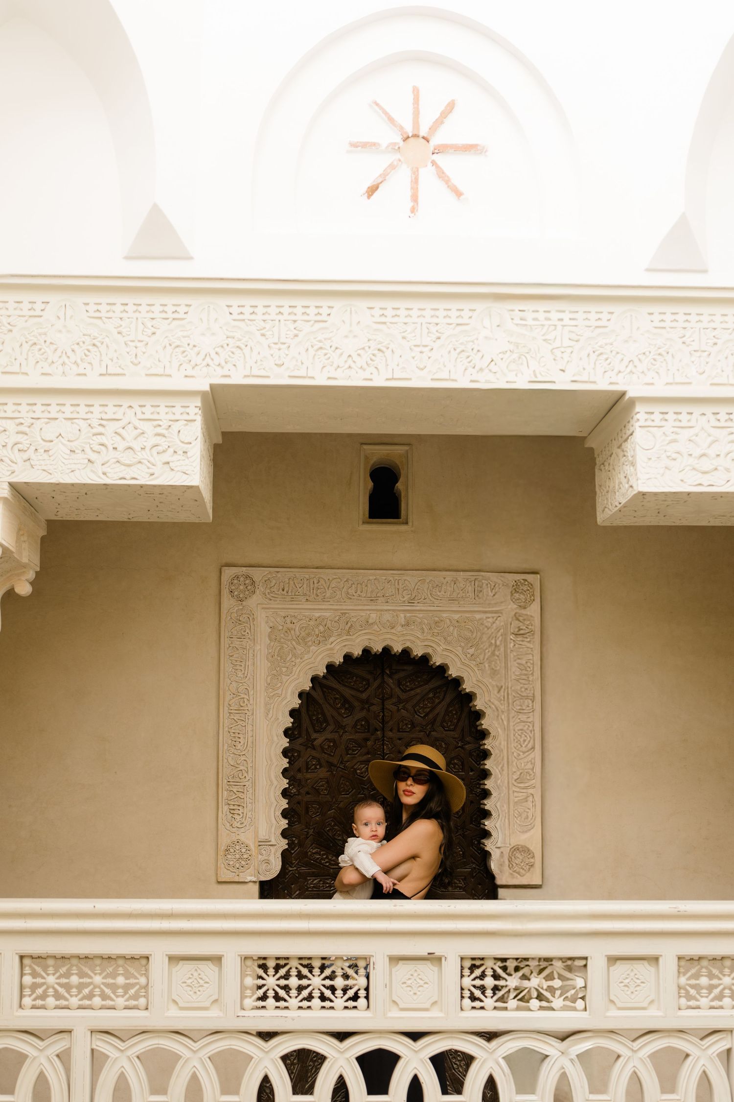 Woman holding baby on ornate Moroccan riad balcony with intricate white plasterwork and arched doorway.