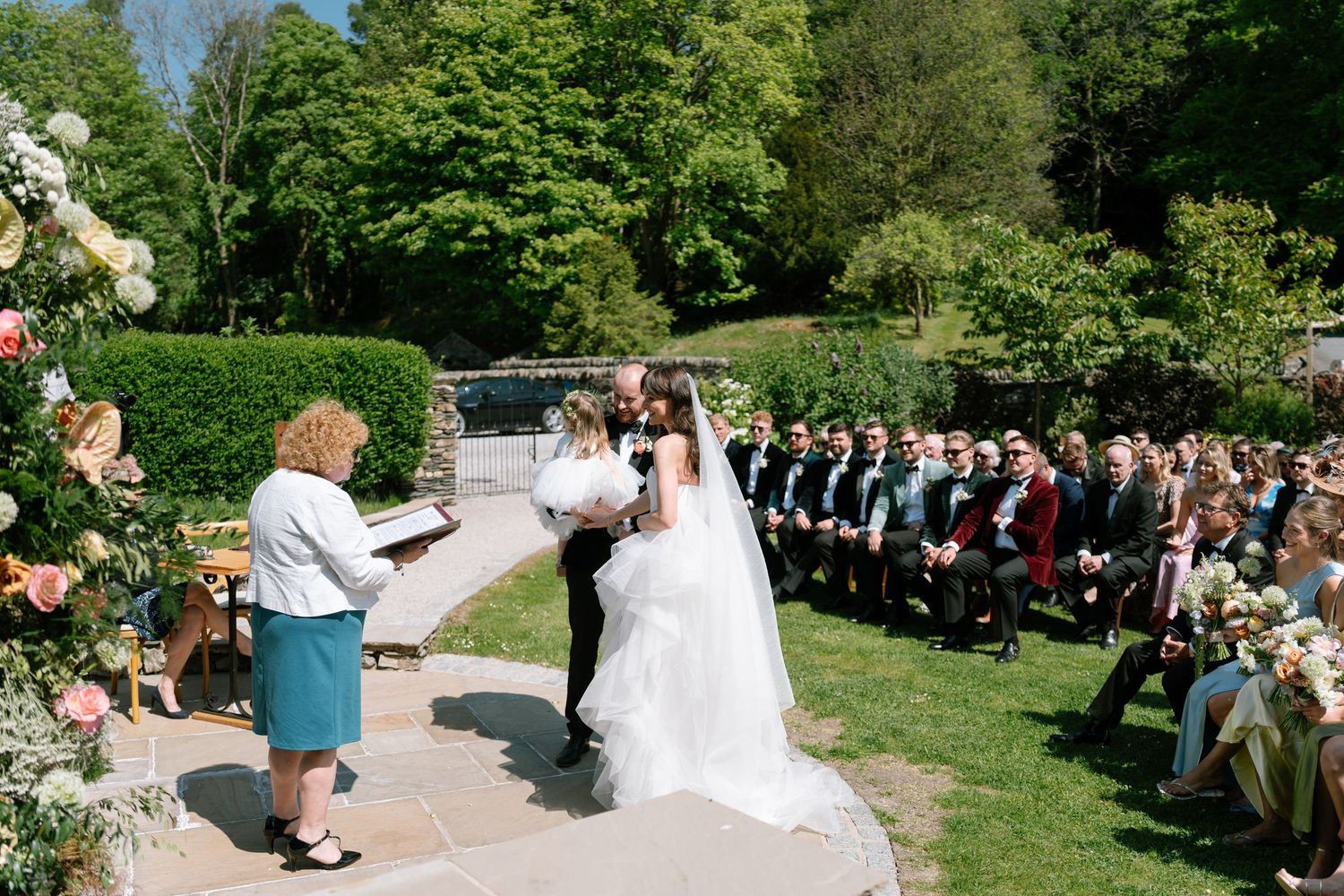 Wedding guests gathered on a sunny lawn with stone buildings in the background during an outdoor ceremony.