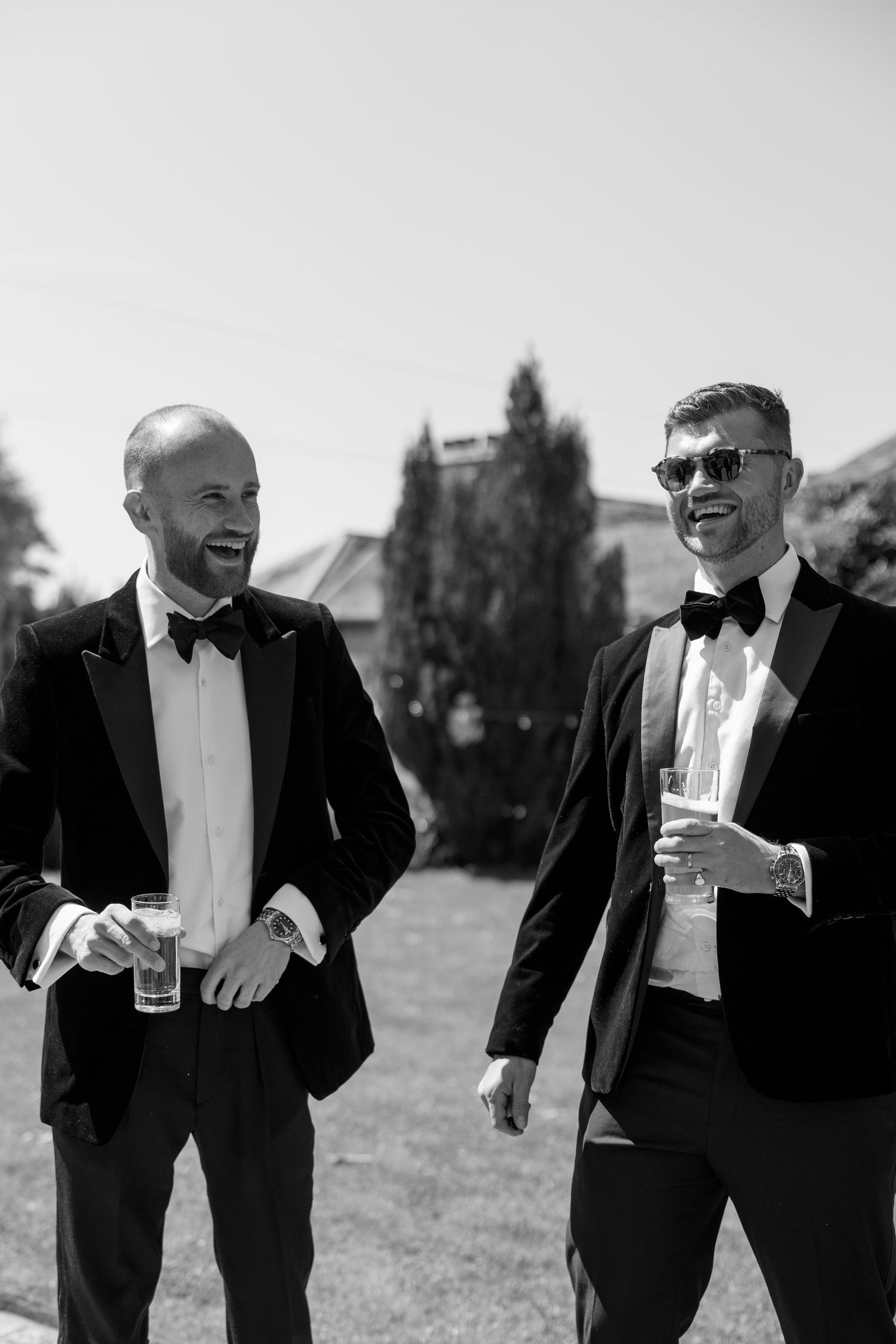 Men in tuxedos and bow ties laughing together at an outdoor event.