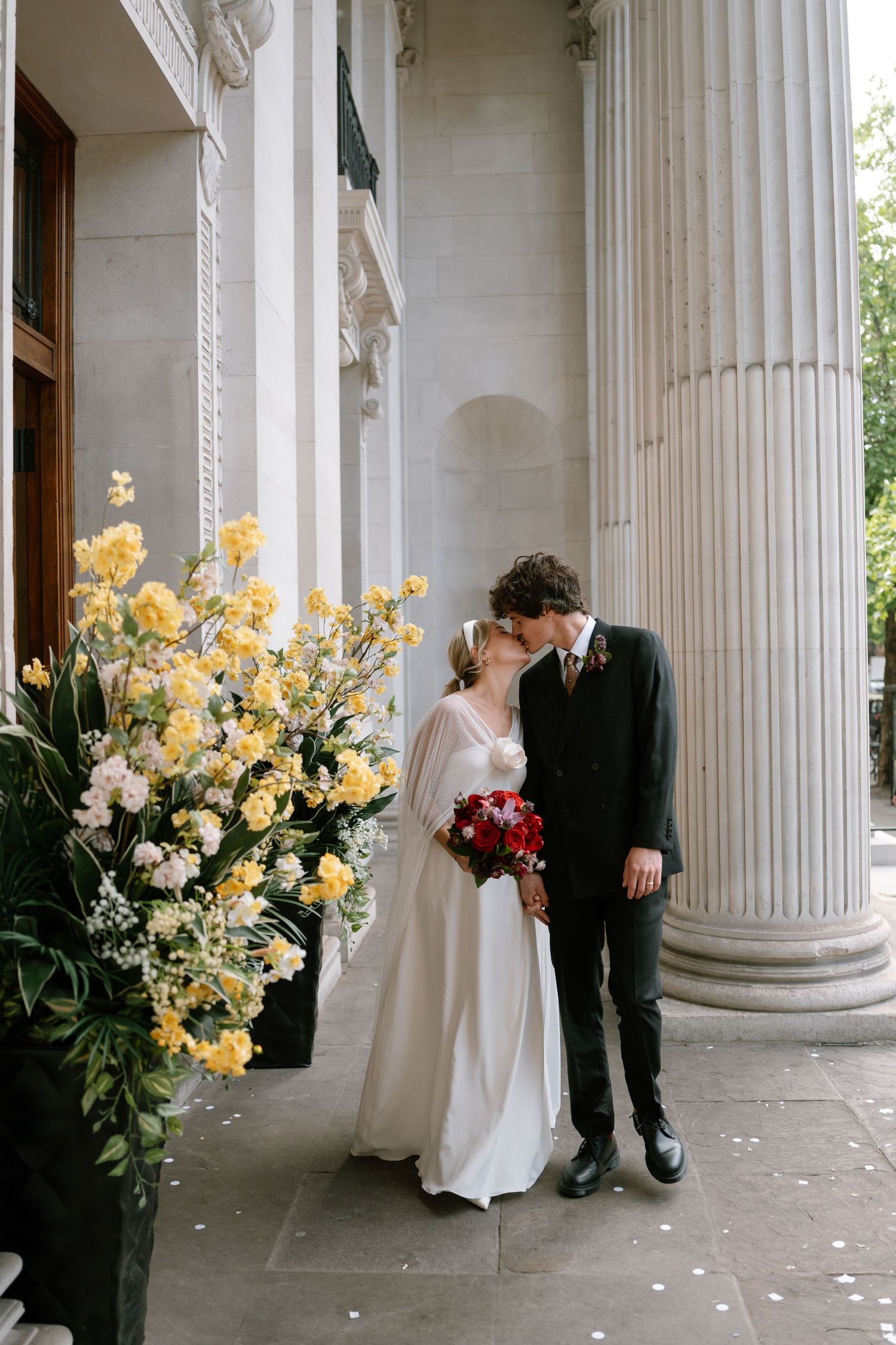 A wedding couple shares an intimate moment between elegant white columns with yellow floral arrangements.