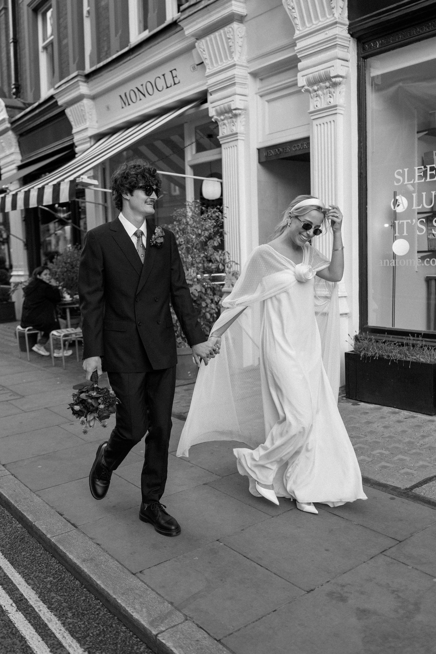 Black and white image of newlyweds walking past classic architecture and shop windows.