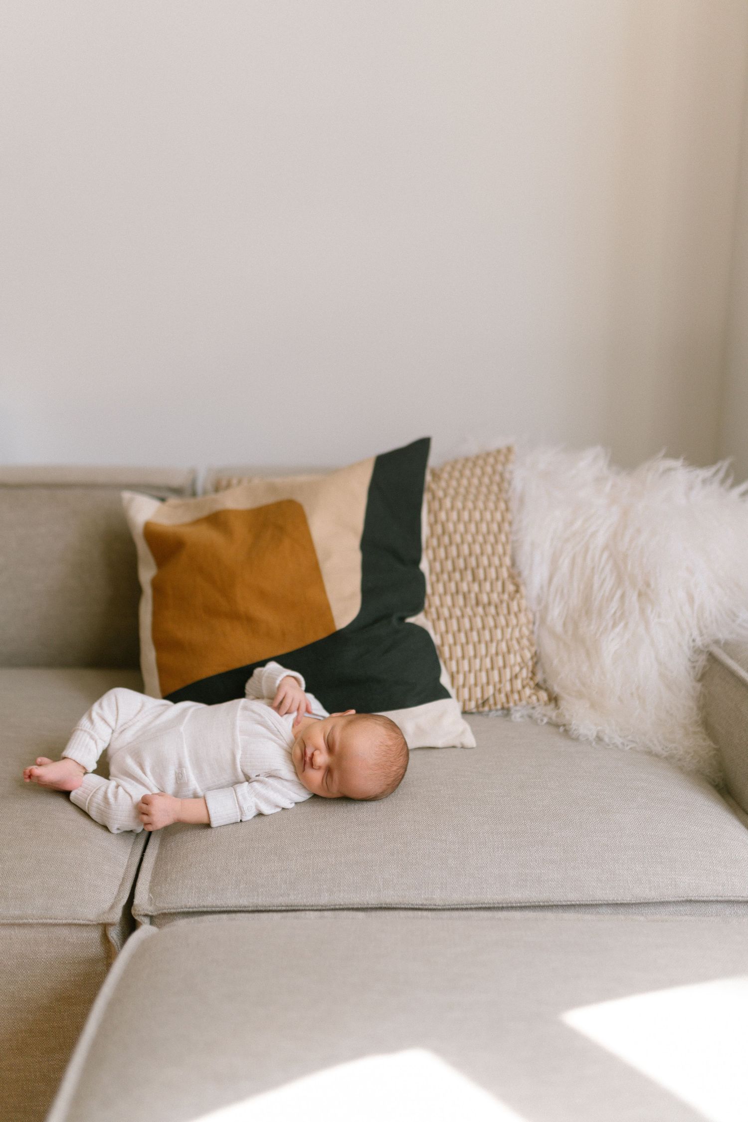 Baby in white clothing sleeping peacefully on a light gray couch with decorative pillows.