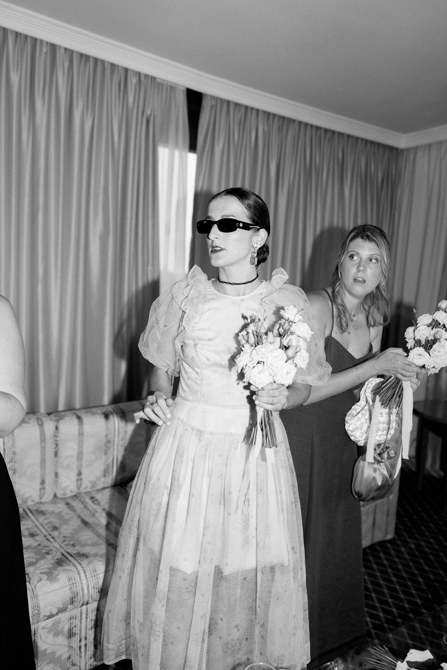 Group wearing formal dresses and holding bouquets in a hotel room.
