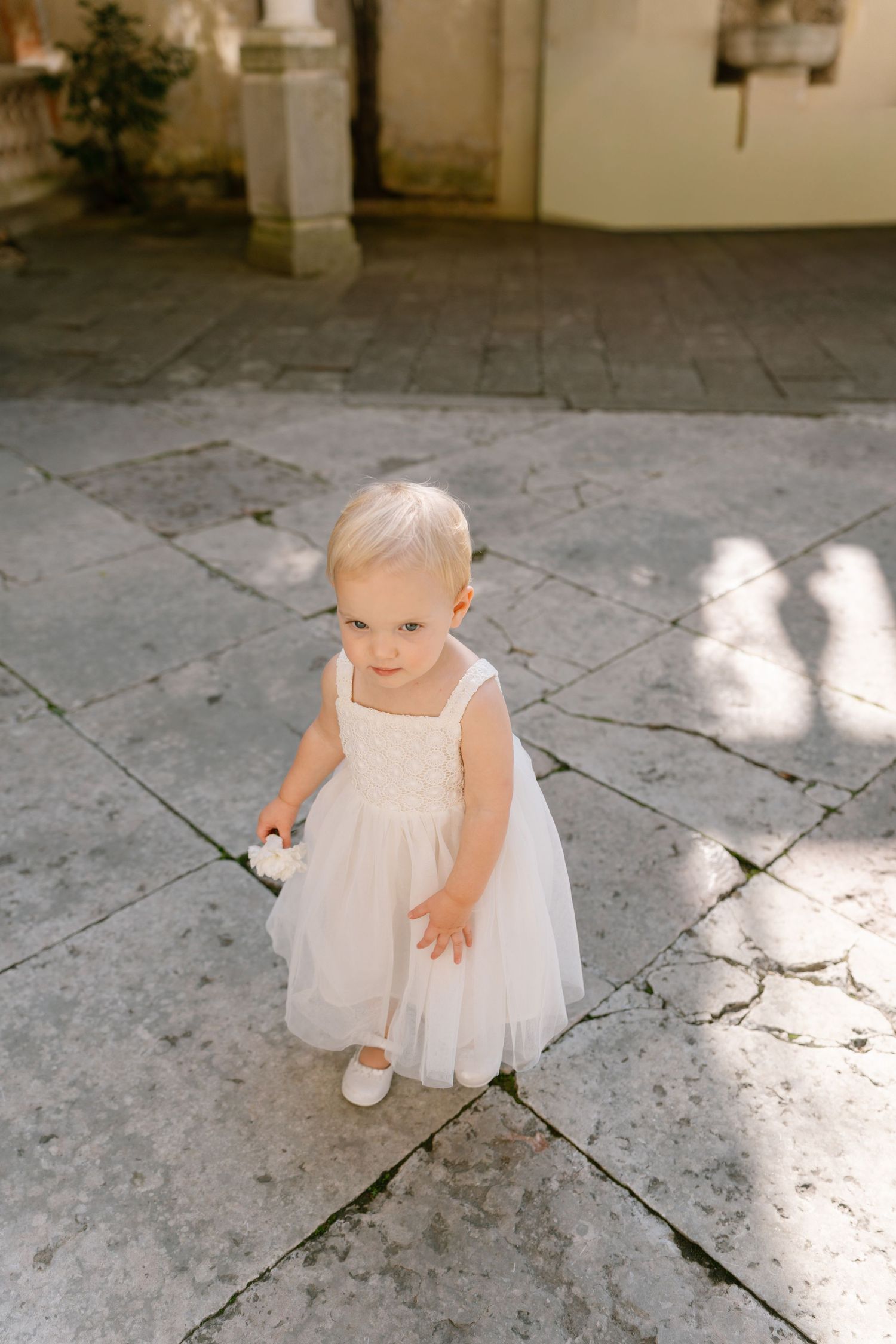Small child in white dress stands on stone courtyard in dappled sunlight.
